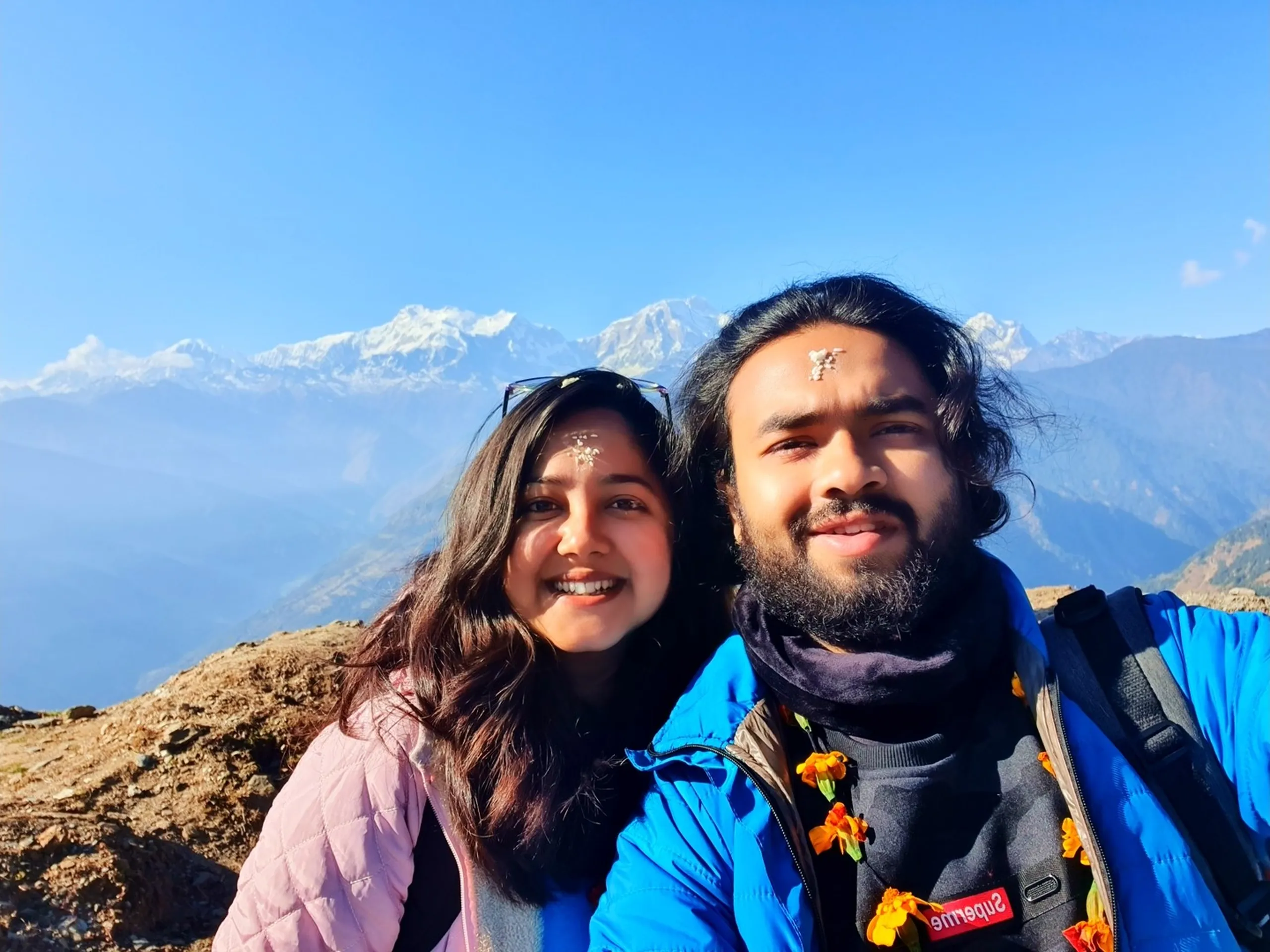 A warm and joyful close-up selfie of two trekkers - a young South Asian woman and a bearded South Asian man - posing together cheek-to-cheek at the Ghale Gaun high-altitude viewpoint in Lamjung District, Nepal, both smiling broadly and radiating the happiness of a successful trek. The woman on the left has long dark wavy hair, wears a light pink quilted puffer jacket, and has a small white rice-flour tika blessing mark on her forehead along with sunglasses perched on top of her head. The man on the right has a full beard and long dark hair, wears a blue puffer jacket over a Supreme black sweatshirt, and also has a white tika on his forehead along with orange marigold garland flowers around his collar. In the background, the magnificent Annapurna Himalayan snowpeaks - Annapurna II and Lamjung Himal - gleam brilliant white against a clear blue sky above hazy blue mountain valleys. Keywords: Nepal trekking selfie couple Annapurna, Ghale Gaun summit selfie tika garland, Annapurna range selfie Nepal, tika forehead Nepal cultural blessing, Gurung village welcome tradition Nepal, Ghale Gaun trek selfie 2019, Nepal Himalayan team friends selfie, Annapurna Conservation Area selfie, marigold garland tika Nepal trekkers, Nepal mountain selfie December 2019.