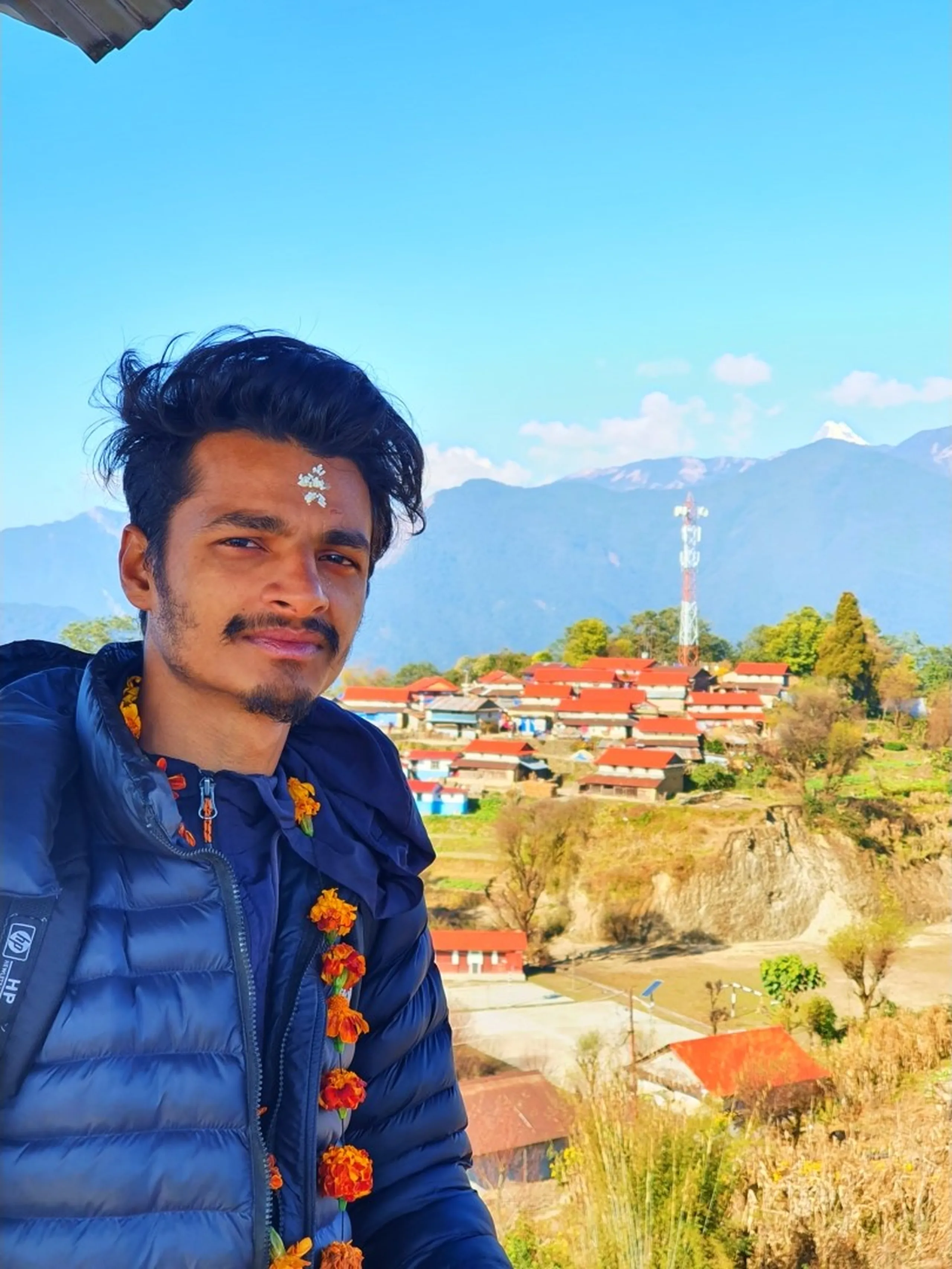 A charming close-up portrait selfie of a young South Asian man in his mid-twenties, wearing a navy blue quilted puffer jacket with a small HP logo on the chest, a traditional orange and red marigold flower garland draped around his neck, and a white rice-flour tika blessing mark on his forehead - marks of a Gurung cultural welcome ceremony at Ghale Gaun. He has short, slightly tousled dark hair and wears a subtle half-smile with a confident sideways glance. Behind him, the full panoramic view of Ghale Gaun village spreads across the mid-ground: clusters of traditional stone and mud-brick houses with bright red corrugated iron rooftops, a red-and-white mobile telecommunications tower, bare winter trees, and small buildings descend the hillside under a brilliant blue sky. In the far upper background, the snow-dusted peaks of the Annapurna Himalayan range and Lamjung Himal are faintly visible above the mountain ridges. Keywords: Ghale Gaun village selfie portrait, marigold garland tika Gurung welcome, Ghale Gaun red roof village Nepal, Lamjung District Nepal village trek portrait, Annapurna Himal behind village Nepal, Nepal cultural welcome trekkers, Gurung village hospitality Nepal, Ghale Gaun mobile tower Nepal, Nepal mountain village portrait December 2019, Annapurna Conservation Area village selfie.