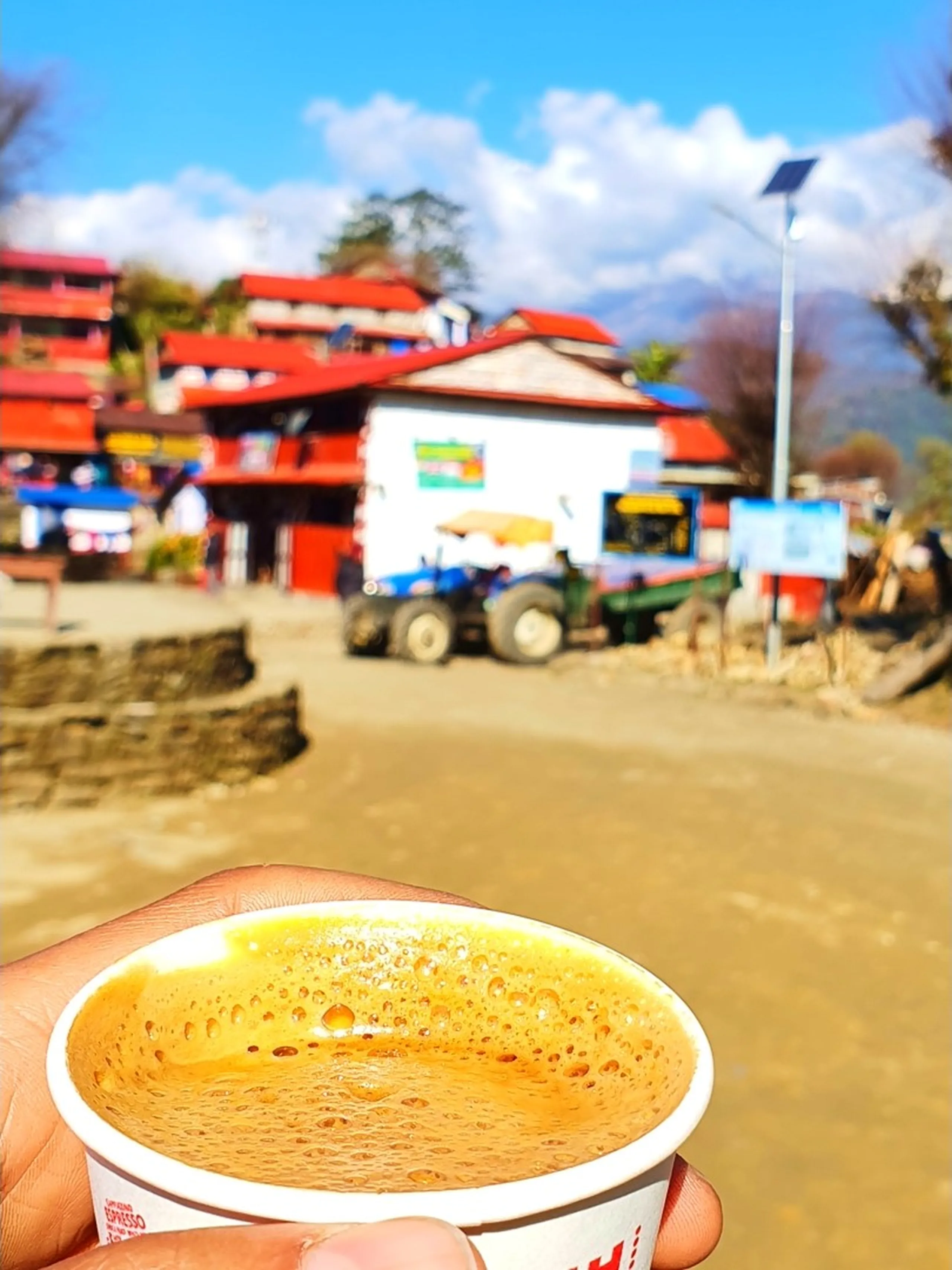 A close-up, intimate food and travel photograph taken at eye level showing a hand holding a small disposable paper cup filled to the brim with steaming hot golden-amber Nepali milk tea (chiya) with a frothy foamy surface studded with tiny bubbles - the classic trekker's comfort drink after a long mountain hike. The cup has printed text 'Espresso' in red lettering. The background is beautifully blurred in soft bokeh, revealing the colorful main street of Ghale Gaun's lower settlement area: red-painted buildings with red corrugated roofs and blue tarpaulin overhangs, a solar street light pole, a blue farm tractor, green vegetation, and various mountain-village commercial signs. A bright blue sky with white clouds is visible above the buildings. The image captures the simple but deeply satisfying joy of a warm cup of Nepali tea in a Himalayan village. Keywords: Nepali chiya mountain tea Ghale Gaun, hot milk tea Nepal trekking, Nepal chiya travel photography, Ghale Gaun village tea cup, trekking tea break Nepal, Lamjung District Gurung village tea, Nepal mountain tea culture, Annapurna Conservation Area village tea, chai Nepal Himalayan trek, Ghale Gaun local tea shop 2019.