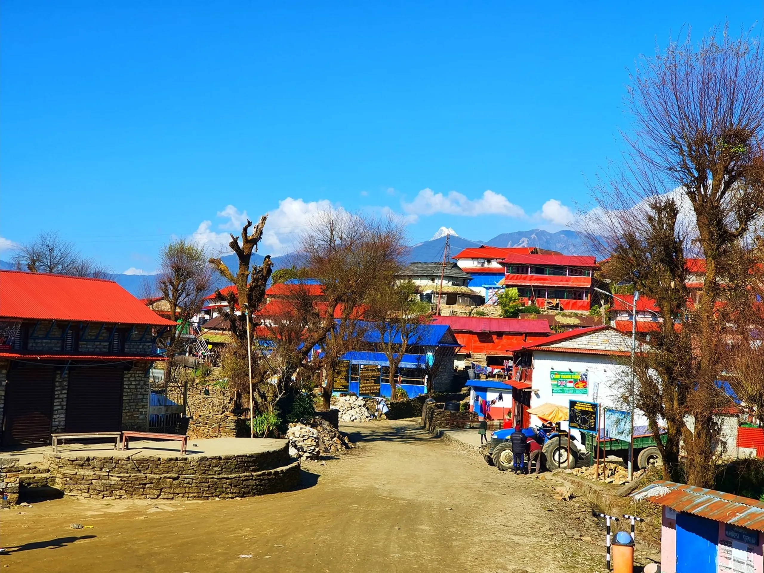 A wide street-level landscape photograph of the main village square and central pathway of Ghale Gaun, the famous Gurung village destination in Lamjung District, Nepal, captured in brilliant midday winter sunshine under a clear blue sky. The scene shows a wide unpaved sandy dirt path running between stone-walled enclosures into the heart of the village. On the left stands a solid stone-built structure with a curved stone-wall bench. Ahead and to the right, multiple multi-storey buildings with vivid red corrugated metal roofs and blue-painted facades line the path - guesthouses, teashops, and local establishments typical of a trekking village. Signboards in Nepali and English are visible, including a 'Babu Trekking Guide' sign in green and white. A blue farm tractor is parked mid-path. Bare winter deciduous trees frame the left and right edges of the frame. In the far background above the rooftops, the distinctive snow-capped conical peak of what appears to be Lamjung Himal or a nearby Annapurna sub-peak pierces the blue sky. Keywords: Ghale Gaun main village street Nepal, Gurung village Lamjung Nepal street view, Ghale Gaun guesthouses red roofs, Nepal mountain trekking village, Lamjung District Gurung village center, Ghale Gaun village square Nepal, Nepal trekking village street photography, Annapurna Conservation Area village, Ghale Gaun Nepal guide signboard, Nepal mountain village daily life 2019.