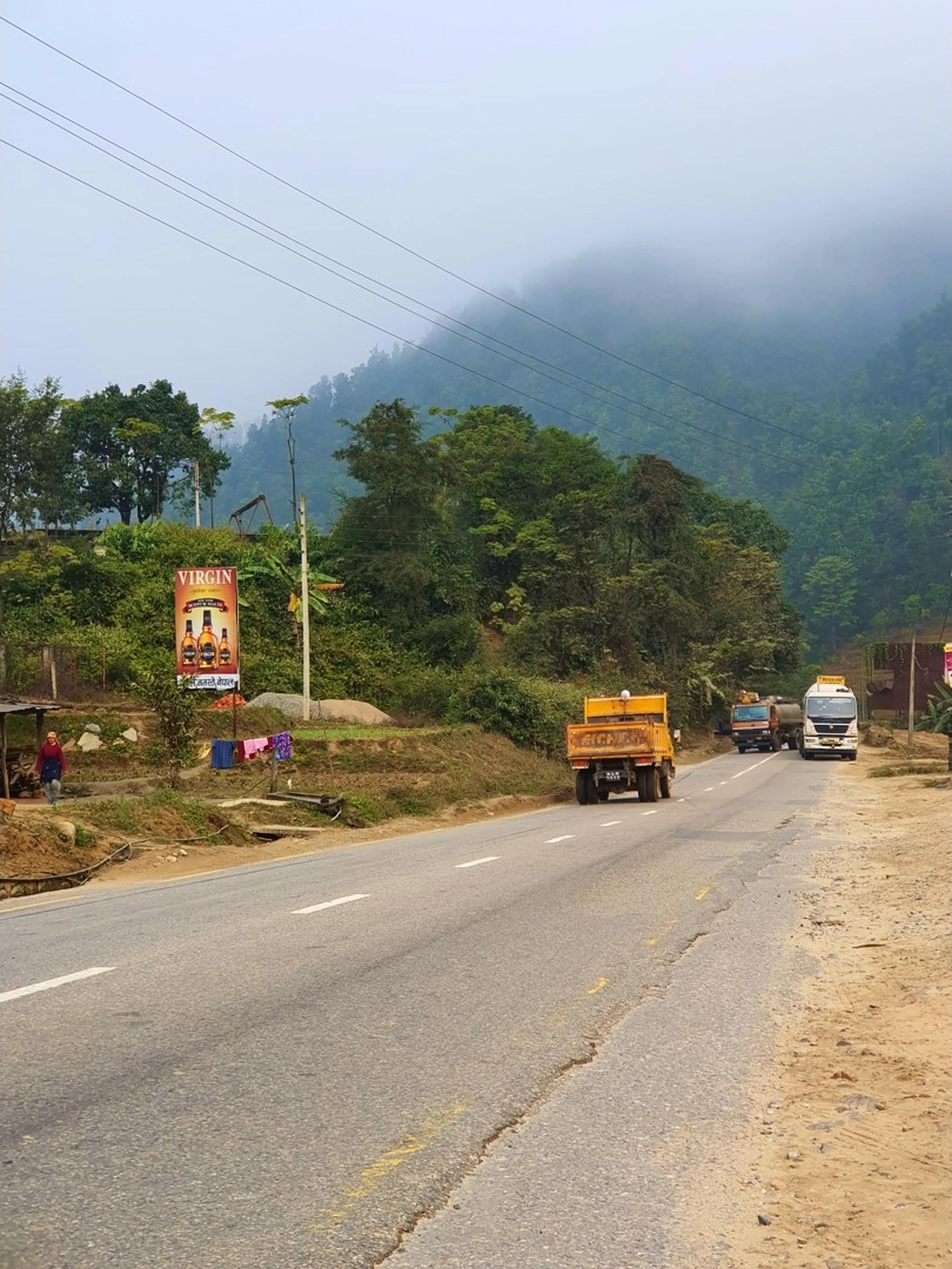 A misty mountain highway in Lamjung District, Nepal, winding through lush green subtropical foliage with cloud-covered forested hills rising steeply in the background under a hazy pale blue sky. A yellow dump truck drives ahead on the two-lane paved road while a tanker truck follows behind. On the left roadside stands a large billboard advertising 'Virgin' whisky bottles, with a small roadside shack nearby, drying laundry hanging on a line, and a lone figure walking along the road edge. The scene captures the raw, unhurried character of a Nepali mountain highway en route to Besisahar and the Annapurna trekking region. Keywords: Lamjung Nepal highway travel, Besisahar road Annapurna trek, Nepal mountain road photography, foggy Nepali highway, road to Ghale Gaun Lamjung, Annapurna Conservation Area road, Nepal travel vlog scenery, trekking route Annapurna region Nepal 2019.