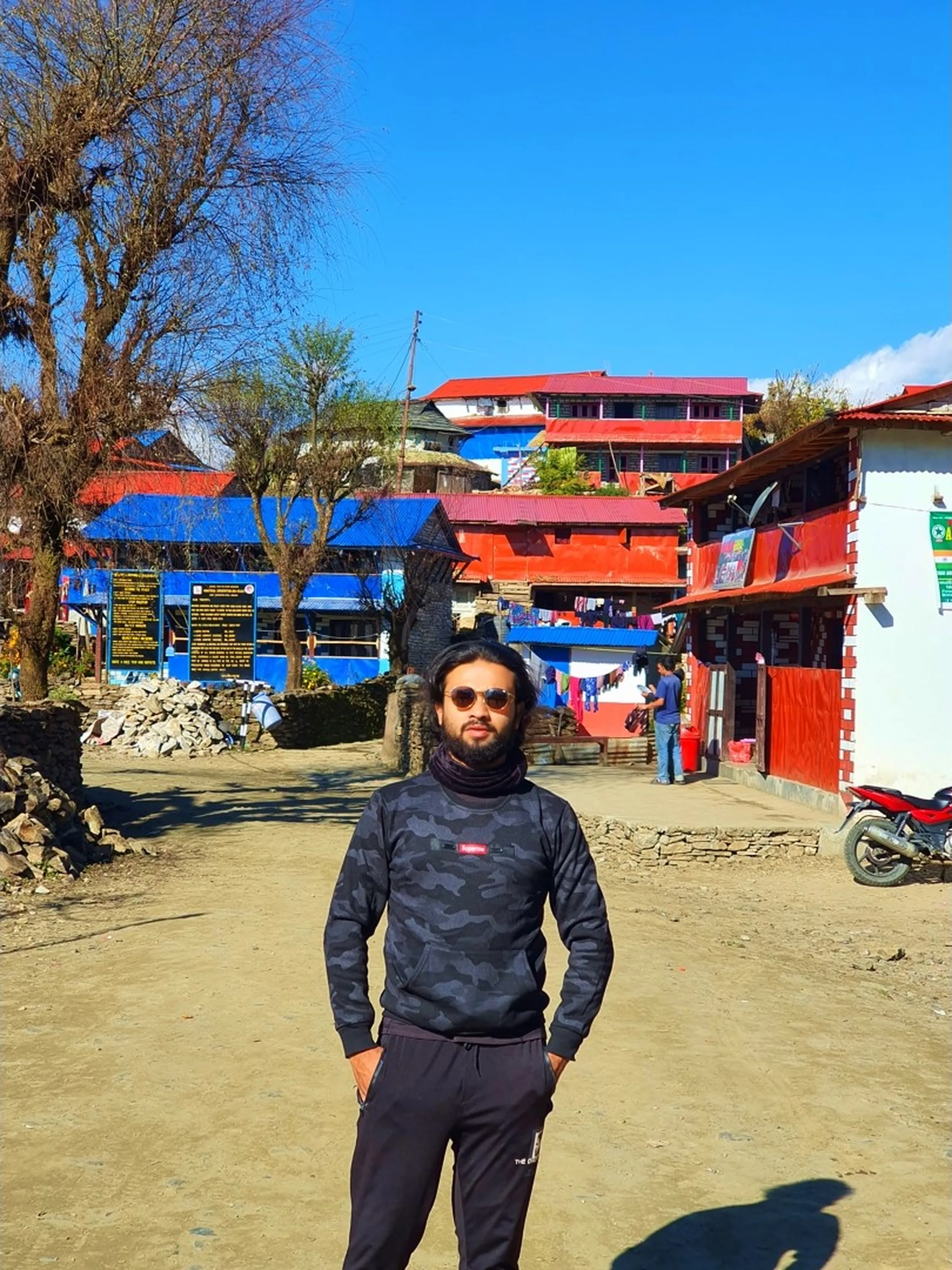 A casual full-body portrait of a bearded South Asian man with long dark hair, wearing a dark grey and black camouflage-pattern Supreme-branded long-sleeve sweatshirt, matching dark track pants from The North Face, round-framed brown-tinted sunglasses, and a dark neck gaiter pulled down, standing relaxed with both hands in his pockets on the sandy unpaved central square of Ghale Gaun village in Lamjung District, Nepal. Behind him, the colorful architecture of the Gurung village unfolds - vivid blue-painted buildings, red corrugated rooftop guesthouses, a local conservation area information board in Nepali and English, stone walls, laundry drying on a line, a red motorcycle parked at the right edge, and a bare tree on the left side under a brilliant cloudless blue sky. This appears to be taken on the return leg of the trek, the man in casual trekking wear exploring the village before heading back. Keywords: Ghale Gaun village portrait Nepal, Supreme sweatshirt Nepal trek style, Gurung village center Lamjung Nepal, Ghale Gaun blue building red roof, Nepal mountain village exploration, Annapurna Conservation Area village portrait, Ghale Gaun village daily life, Nepal trekking style casual portrait, Lamjung District Gurung village trek 2019, Nepal cultural village trekking experience.