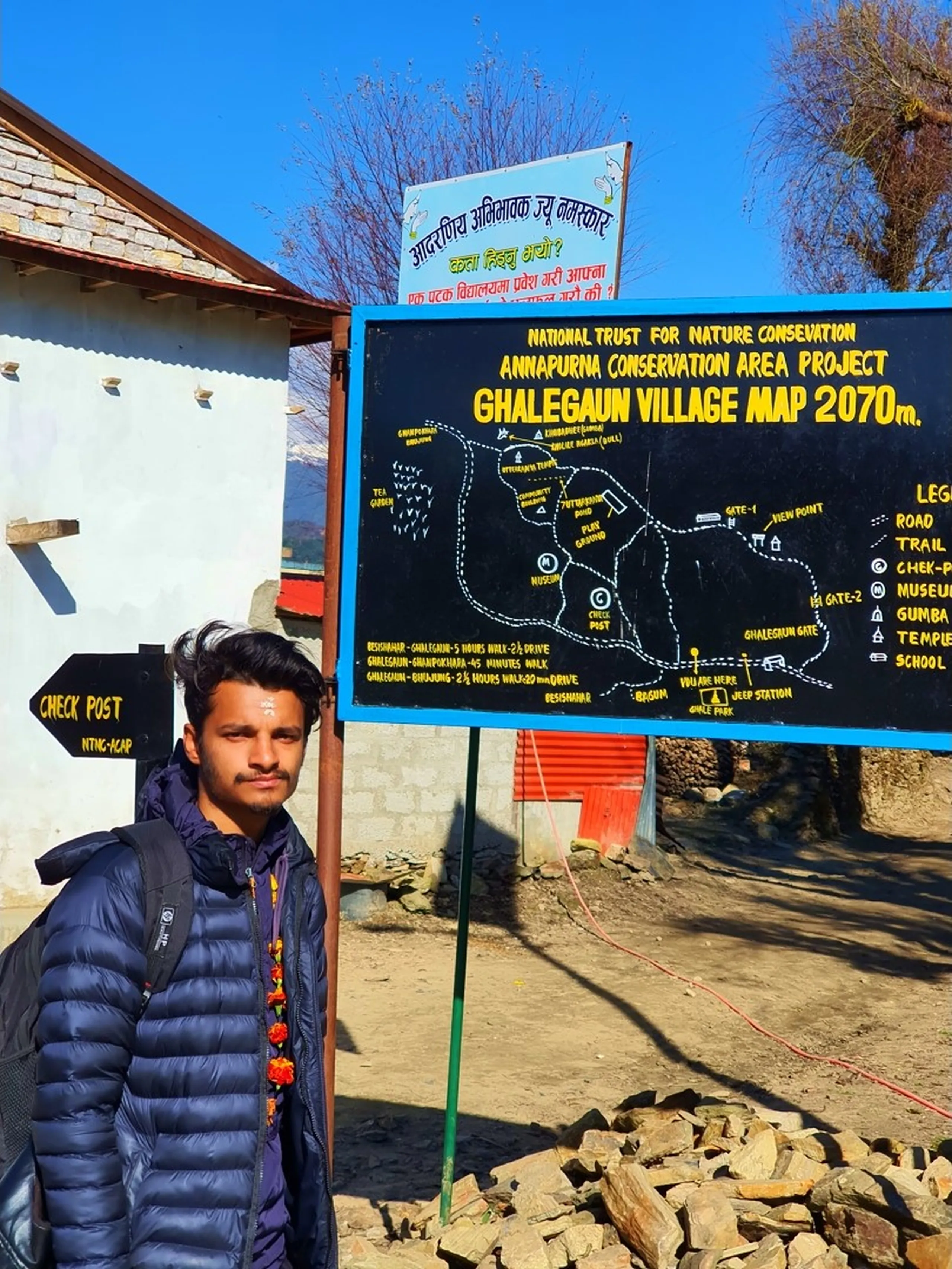 A young Nepali male trekker wearing a navy blue puffer jacket and backpack adorned with a marigold garland stands beside the official Ghalegaun Village Map signboard at 2070m elevation, installed by the National Trust for Nature Conservation under the Annapurna Conservation Area Project (ACAP). The large black and yellow map board displays trail routes, checkpost locations, tea gardens, community forest, museum, viewpoint, Ghumpokharajhara, and distance information to Besisahar and Bhujung. A smaller black wooden signpost pointing left reads 'CHECK POST - NTNG-ACAP'. Traditional stone walls, a white plastered building, and a clear bright blue winter sky complete the frame. Shot in crisp morning light in December 2019, this is the official entry point to Ghale Gaun, a popular Gurung village trekking destination in Lamjung District, Nepal - Annapurna Conservation Area trekking, Ghale Gaun trek Nepal, Lamjung district hiking, ACAP checkpost Nepal, Gurung village Nepal trek.