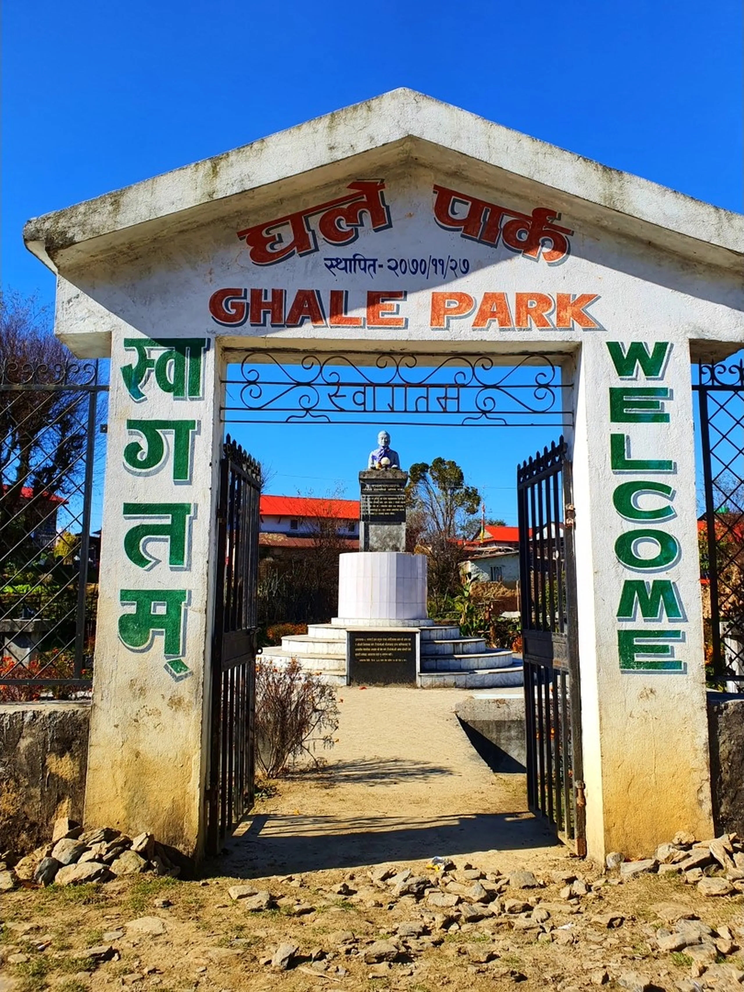 The ornate entrance gate of Ghale Park (??? ?????) in Ghale Gaun village, Lamjung, Nepal, photographed under a vivid deep blue winter sky. The white painted concrete arch gateway is inscribed with 'GHALE PARK' in bold orange letters, with the Nepali script '??? ?????' above and an establishment date '????/??/??' (2070 BS). Green Devanagari letters spelling '????????' (Welcome) run vertically down the left pillar, and the English word 'WELCOME' runs vertically in bold green on the right. Black wrought iron gates stand open, revealing a straight stone-paved path leading to a white pedestal monument or bust statue at the center of the park, surrounded by bare-branched trees, a red-roofed building, and dry winter garden beds. Rocky ground and remnant stone rubble edge the foreground - Ghale Gaun village Nepal, Gurung cultural park Lamjung, Annapurna Conservation Area village, Nepal heritage site, Lamjung trekking destination, Nepal rural tourism.