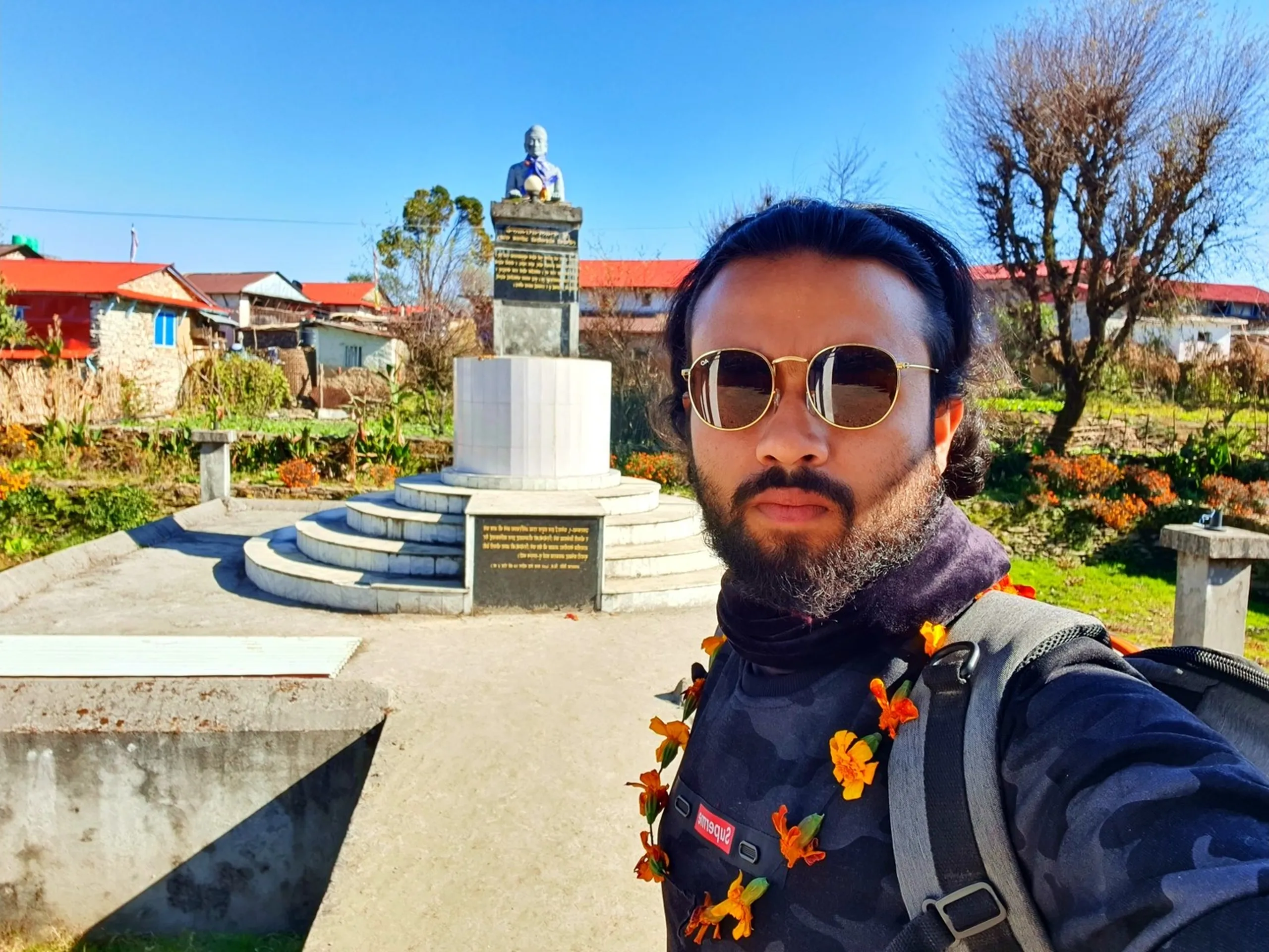 A bearded male trekker with long dark hair tied back takes a selfie inside Ghale Park in Ghale Gaun, Lamjung, Nepal. He wears round gold-rimmed sunglasses, a dark camouflage-pattern jacket with a red Supreme logo label, and carries a gray backpack. A string of vibrant orange marigold flowers - the traditional Nepali 'swagat mala' welcome garland - is draped around his neck and over his pack strap, indicating a warm village welcome. Behind him, a white cylindrical pedestal supports a bust monument statue, with steps and a commemorative plaque with Nepali text below. Red and orange marigold garden beds, bare winter trees, colorful village homes with red and tin roofs, and a crystal-clear deep blue Himalayan sky fill the background - Ghale Gaun Nepal trekking, Gurung village welcome garland Nepal, Annapurna Conservation Area winter trek, Lamjung Nepal adventure travel, Nepal cultural trekking experience, marigold garland Nepal tradition.