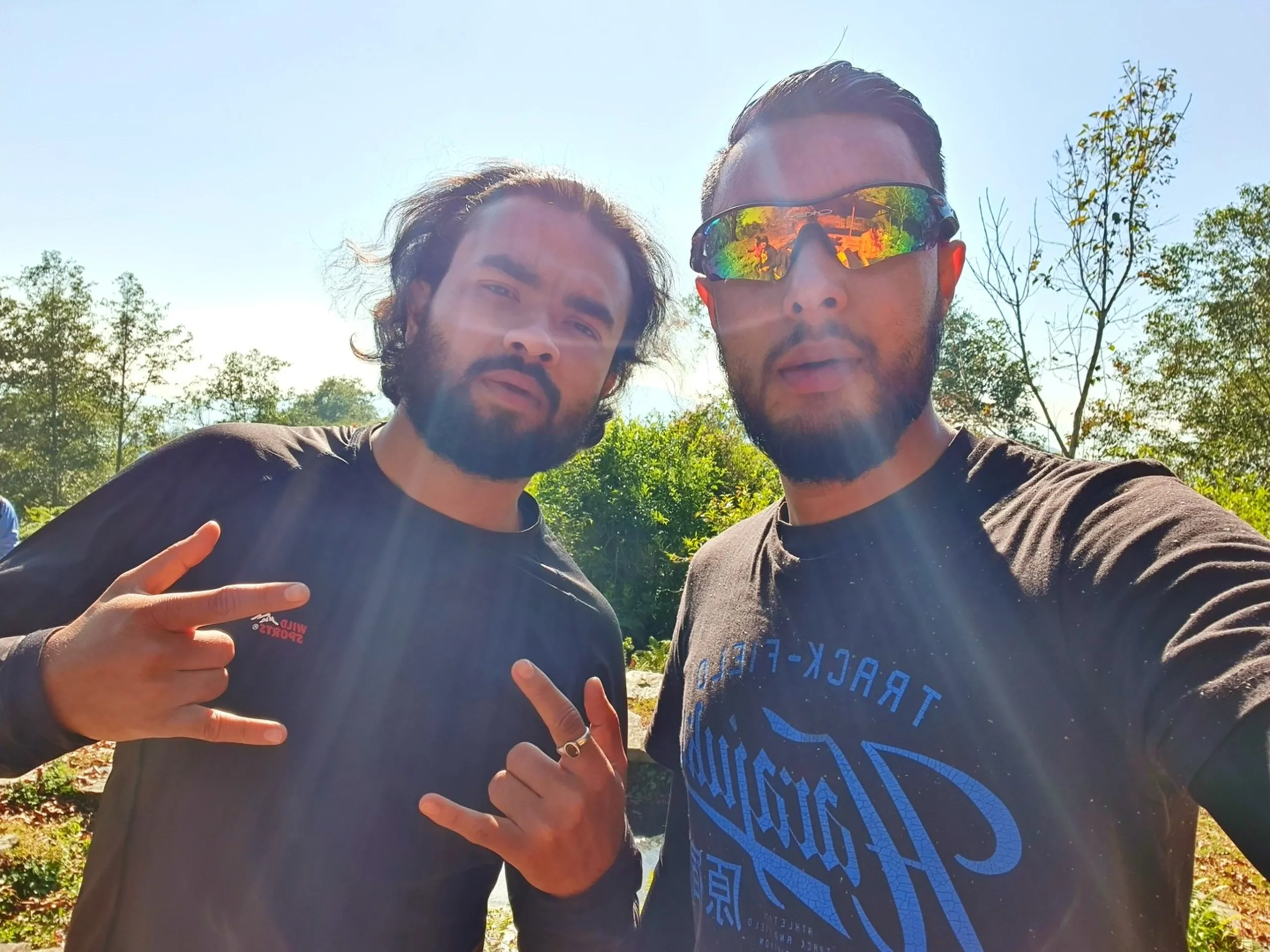 Two young Nepali male trekkers pose for a close-up selfie outdoors against a sunlit backdrop of lush green trees and bright blue sky. The man on the left has wavy dark shoulder-length hair, a short beard, and wears a black full-sleeve shirt with a small 'Wild Sports' logo; he flashes a double shaka / rock-on hand gesture. The man on the right has short slicked-back dark hair, a trimmed beard, and sports striking iridescent multi-color reflective wraparound sunglasses; he wears a dark black 'Track-Field' graphic T-shirt. Both look directly into the camera with cool, relaxed energy - the embodiment of camaraderie on a corporate group trek. Bright natural sunlight creates lens flare effects - Ghale Gaun group trek Nepal, friends trekking Nepal mountains, Annapurna region adventure group, corporate team trekking Nepal, Lamjung Nepal outdoor adventure, Nepal hiking group selfie.