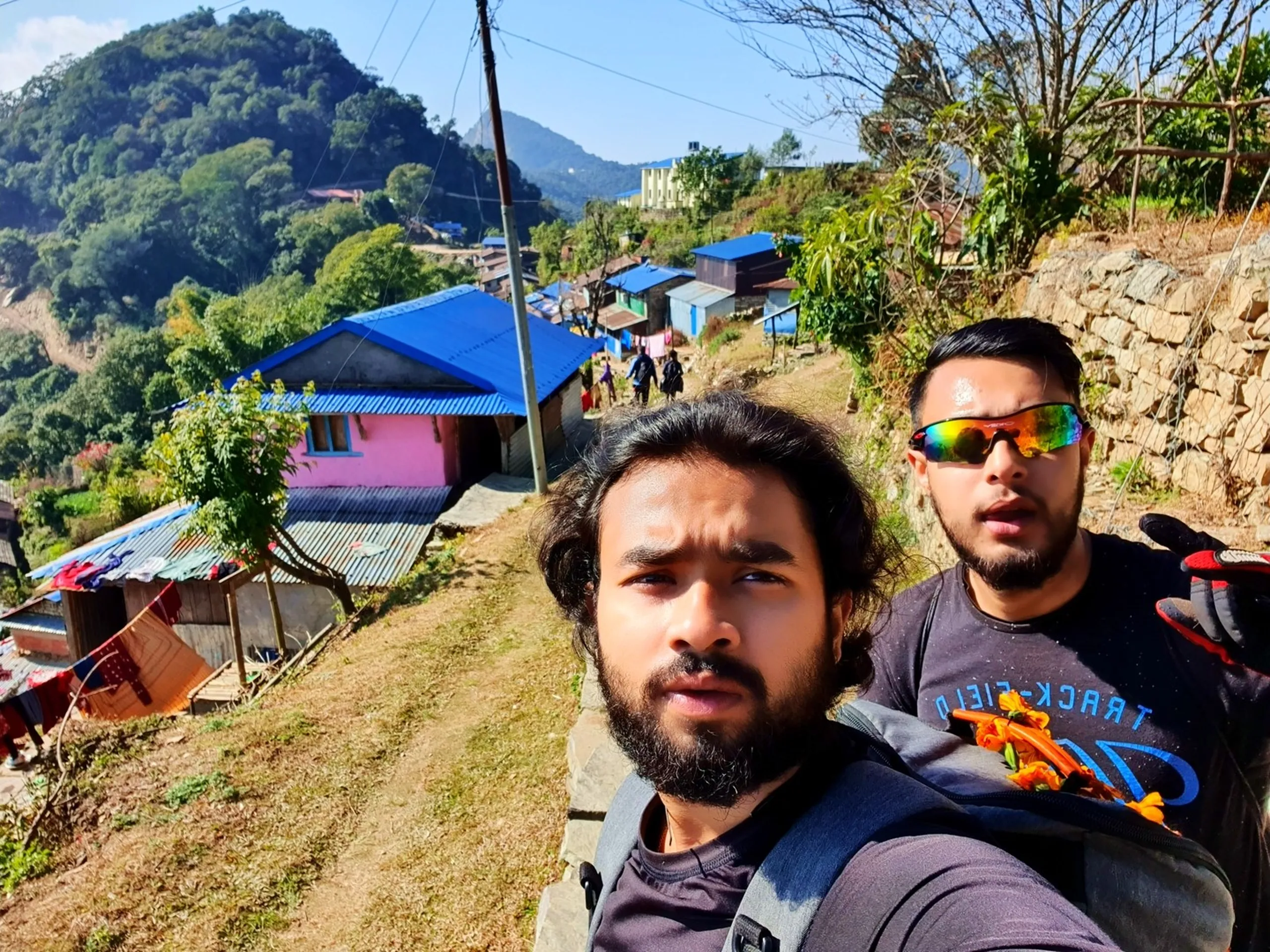 Two bearded male trekkers take a selfie along a narrow mountain ridge trail path in Ghale Gaun village, Lamjung, Nepal. The man in the foreground has long wavy dark hair and wears a dark T-shirt and backpack; behind him, a second trekker in a black 'Track-Field' graphic T-shirt with iridescent sport sunglasses and trekking gloves stands close. In the background, the hillside village of Ghale Gaun cascades downhill, with closely packed houses featuring colorful blue metallic rooftops, corrugated tin, and pink-painted walls. Clotheslines with laundry are visible between homes. Green terraced hillsides and forested ridgelines lead upward to more mountain slopes in the distance under a hazy sunny blue sky. Other trekking group members can be spotted farther along the trail - Ghale Gaun village Nepal, Gurung village trekking Nepal, Lamjung mountain village, Annapurna Conservation Area village tour, Nepal rural mountain community, traditional Nepal hill village homes, group trekking Lamjung Nepal.