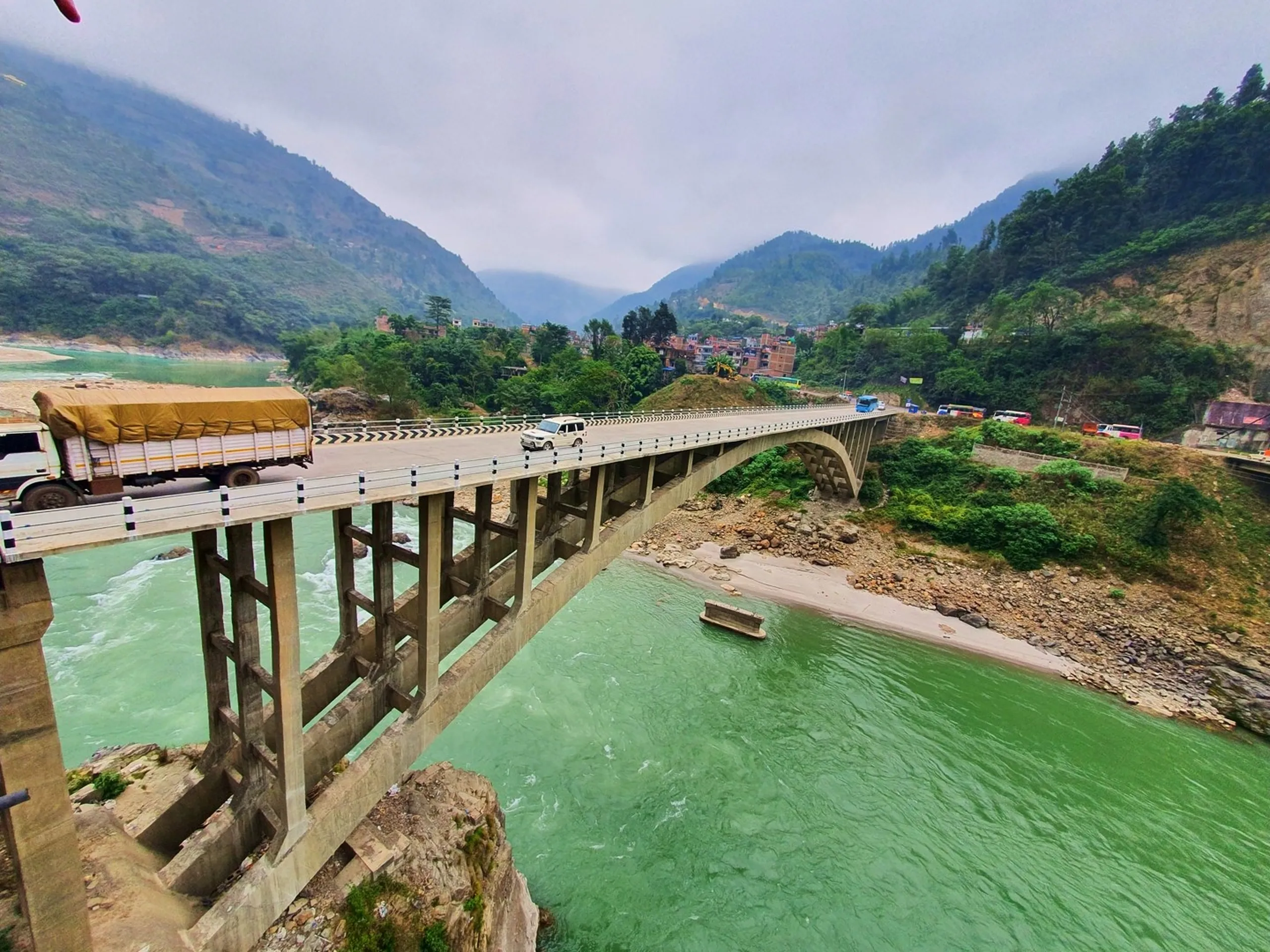 A dramatic wide-angle view of a large concrete arch bridge spanning a rushing turquoise-green glacial river - likely the Marsyangdi River - deep in the Himalayan foothills of Lamjung District, Nepal, on the route to Ghale Gaun and the Annapurna region. A heavy goods truck with an orange tarpaulin cover and a white SUV cross the bridge simultaneously, while lush green forested mountain slopes rise steeply on both sides under a cloudy overcast sky. A small riverside settlement with colorful buildings clings to the hillside on the far bank, and rocky riverbeds flank the emerald water below. The bridge's structural columns are visible from a low-angle perspective, emphasizing its impressive engineering scale. Keywords: Marsyangdi River bridge Nepal, Annapurna trek river crossing, Lamjung District Nepal bridge, turquoise river Nepal mountains, Ghale Gaun approach route, Nepal Himalayan river landscape, Annapurna Conservation Area trek route, Nepal trekking infrastructure, mountain bridge Nepal travel photography 2019.
