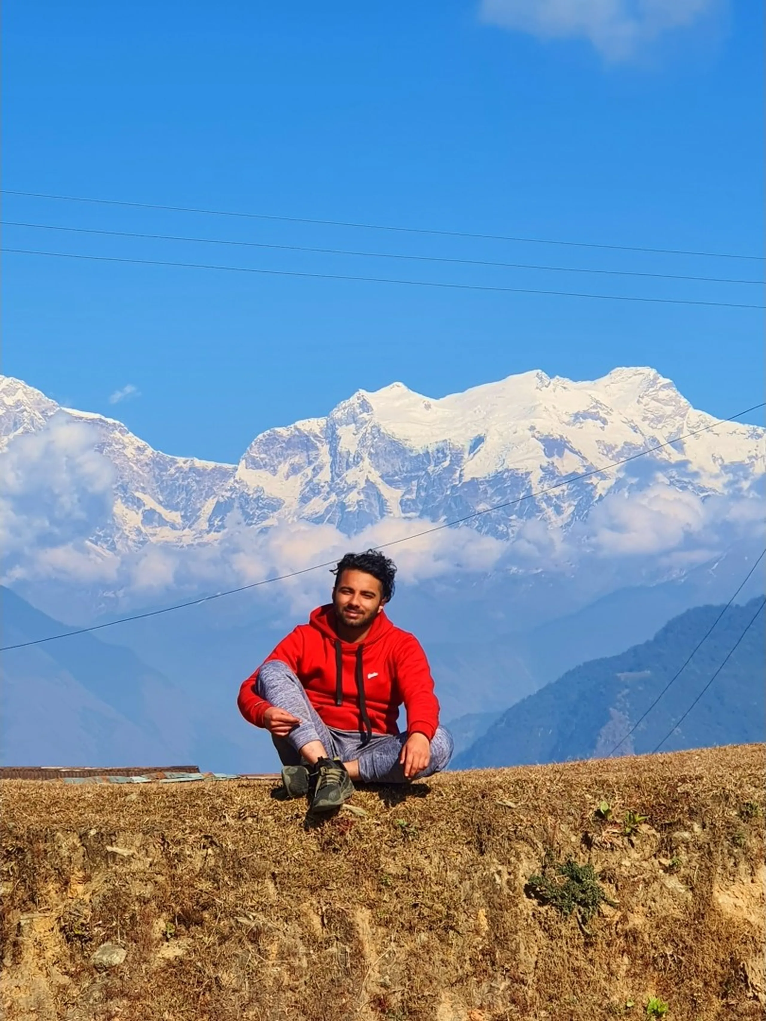 A young Nepali male trekker dressed in a bright red hoodie and light gray jogger pants sits cross-legged on a low earthen terrace wall at an open viewpoint in Ghale Gaun or the surrounding Annapurna Conservation Area, Lamjung, Nepal, smiling at the camera. Behind him, dominating the entire upper half of the frame, is a spectacular panoramic view of the snow-capped Annapurna Himalayan mountain range - massive, multi-peaked glaciated ridgelines of white and blue-gray rising dramatically above a layer of light cloud against a deep cobalt blue winter sky. Two thin power lines cross horizontally in front of the mountains. The foreground shows dry golden-brown grass on the terrace ledge - Annapurna range viewpoint Nepal, Ghale Gaun Himalaya view, Lamjung Annapurna panorama, snow-capped Himalaya Nepal trekking, Annapurna Conservation Area mountain view, best Himalaya viewpoint Nepal, Nepal winter trek Annapurna, Gurung village mountain panorama, Nepal travel bucket list view.
