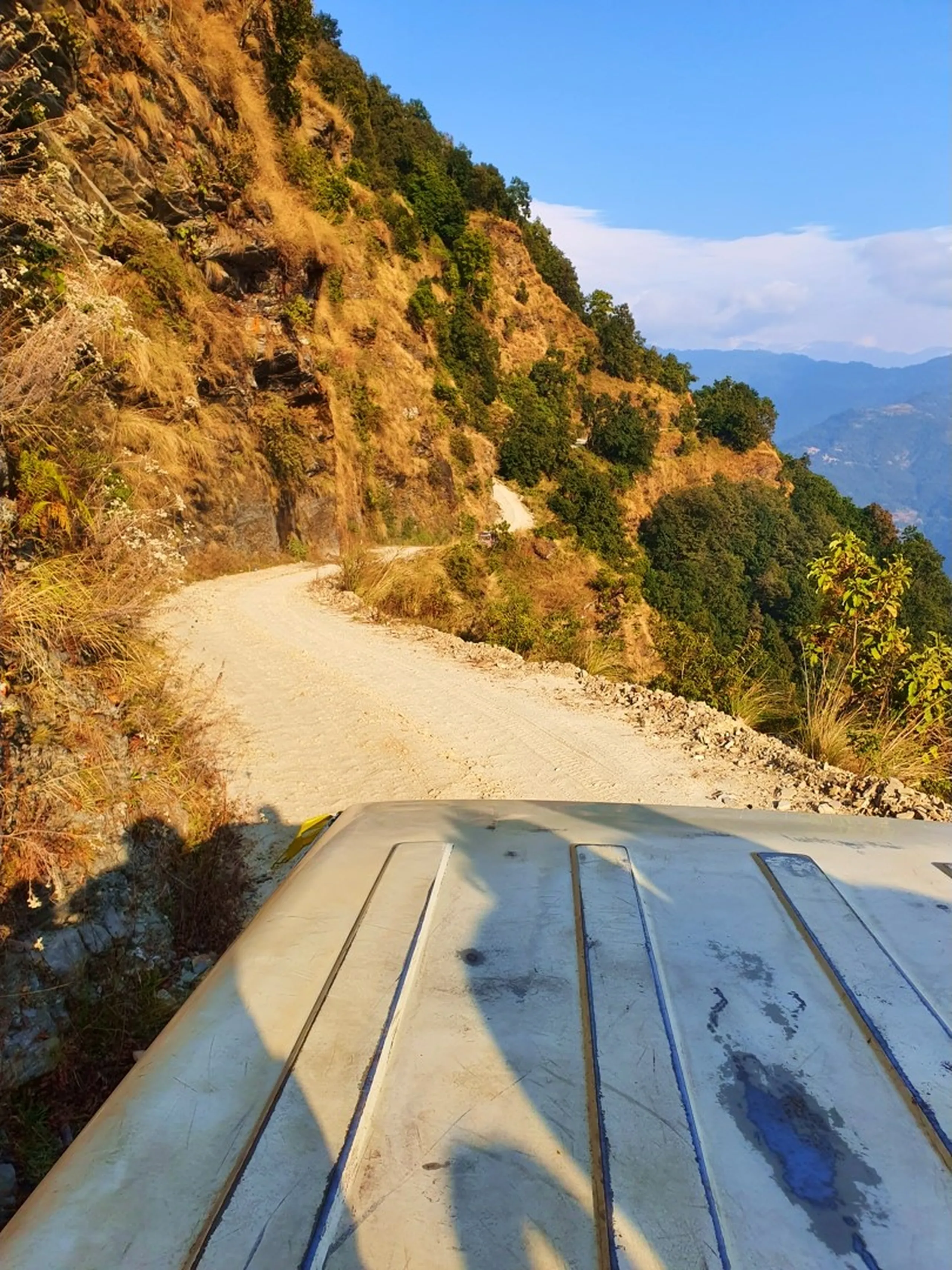A thrilling first-person perspective from the roof of a local Nepali jeep or vehicle traveling along an extremely narrow, unpaved mountain road carved into a steep sun-drenched rocky cliff face in the Annapurna foothills of Lamjung, Nepal - on the adventurous ascent toward Ghale Gaun. The dusty white gravel track hugs the sheer orange-brown mountainside with no guardrails, curving sharply around a blind bend. Dry golden grass and scattered green bushes cling to the rocky slope above, while the road drops off steeply to the right revealing distant blue mountain ridges and valleys far below bathed in bright afternoon sunlight. The white ribbed rooftop of the vehicle occupies the lower foreground of the frame. Keywords: Nepal mountain road jeep ride, Ghale Gaun approach road, narrow cliff road Nepal Lamjung, off-road mountain Nepal adventure, unpaved Himalayan road trekking, Annapurna foothills dirt road, Nepal adventure travel photography, cliffside road Nepal jeep, dangerous mountain road Nepal trek 2019.