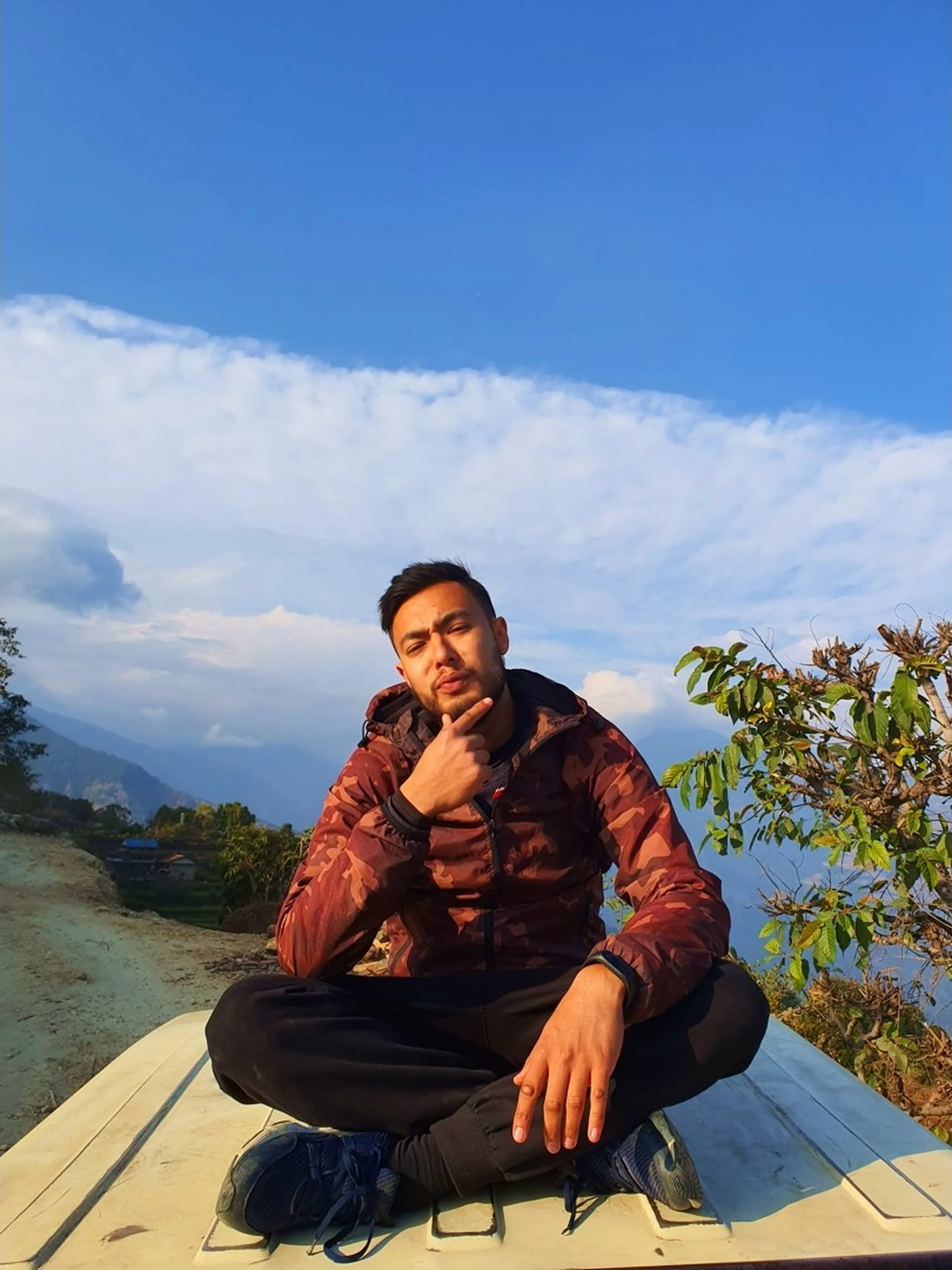 A young South Asian man in his late twenties sits cross-legged in a relaxed, confident pose atop the flat white roof of a moving jeep on a dusty mountain road in the Annapurna foothills near Ghale Gaun, Lamjung District, Nepal. He wears a maroon camouflage puffer jacket, black track pants, and blue sneakers, with one hand resting on his knee and the other raised in a thoughtful chin-touching pose. Behind him stretches a vast panoramic sky of crisp blue with dramatic white clouds hovering over distant dark mountain ridges and green hillside valleys. Dry tree branches are visible to his right. The golden afternoon light sets a warm adventurous tone, perfectly capturing the carefree spirit of trekking in Nepal. Keywords: jeep roof riding Nepal mountains, Ghale Gaun trek adventure, Nepal trekking lifestyle photography, Annapurna region jeep ride, young trekker Nepal Himalaya, adventure travel Nepal 2019, Lamjung District mountain road, Nepal travel blog portrait photography, rooftop jeep ride Nepal.