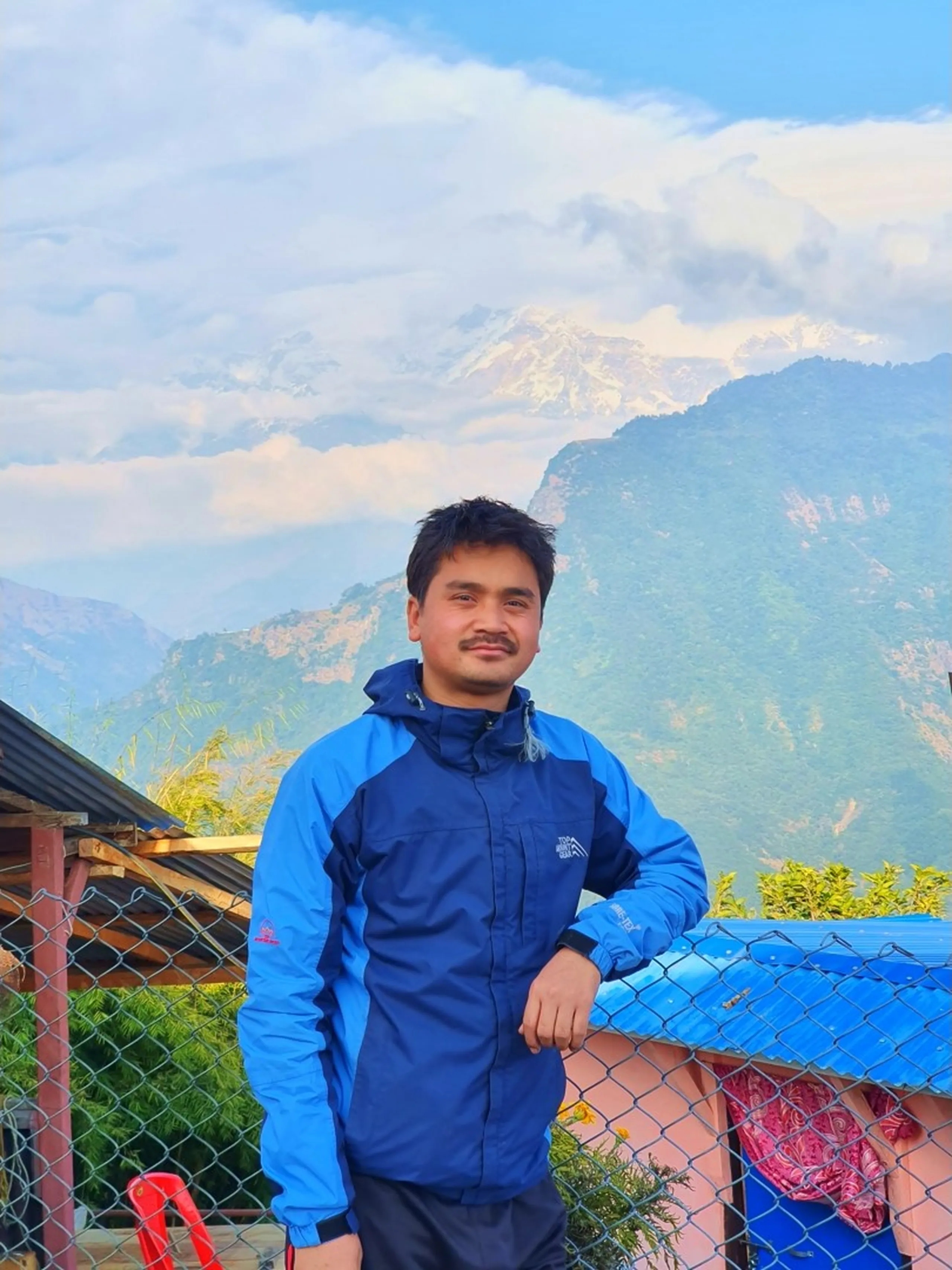 A cheerful young South Asian man in his late twenties, wearing a navy blue two-tone waterproof trekking jacket with a hood, leans casually with one arm resting on a chain-link fence, smiling warmly at the camera. Behind him, a dramatic Himalayan landscape unfolds - layered green forested mountain slopes rise beneath a partially cloudy blue sky, and in the upper background, the brilliant snow-covered peaks of the Annapurna Himalayan range glow white against patches of cloud. A corrugated metal roof, green shrubs, a blue plastic rooftop, and a red plastic chair are visible in the lower background, suggesting a rustic mountain homestay or guesthouse setting in Ghale Gaun, Gurung village, Lamjung District, Nepal. Keywords: Ghale Gaun homestay Nepal, Annapurna snow peak background portrait, Gurung village Nepal trek, Lamjung District trekking guesthouse, Annapurna Conservation Area viewpoint, Nepal mountain portrait photography, waterproof trekking jacket Nepal, smiling trekker Nepal Himalaya, Nepal corporate trek December 2019.