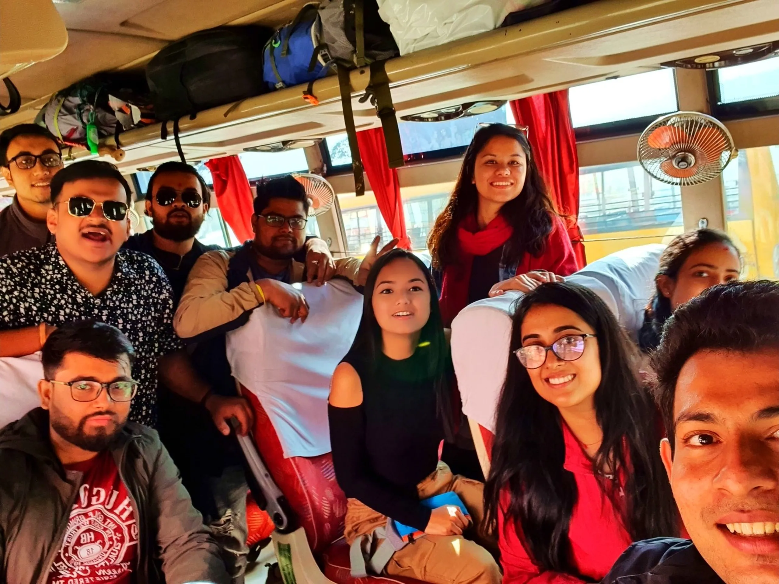 A lively group of young Nepali trekkers taking a wide-angle selfie inside a packed tourist bus headed to Manang, Nepal, with backpacks and trekking gear stuffed in overhead luggage racks, red curtains on the windows, and a ceiling fan visible - capturing the excitement and camaraderie of a Nepal Himalaya group trek from Kathmandu, the kind of squad energy that defines adventure travel in Nepal