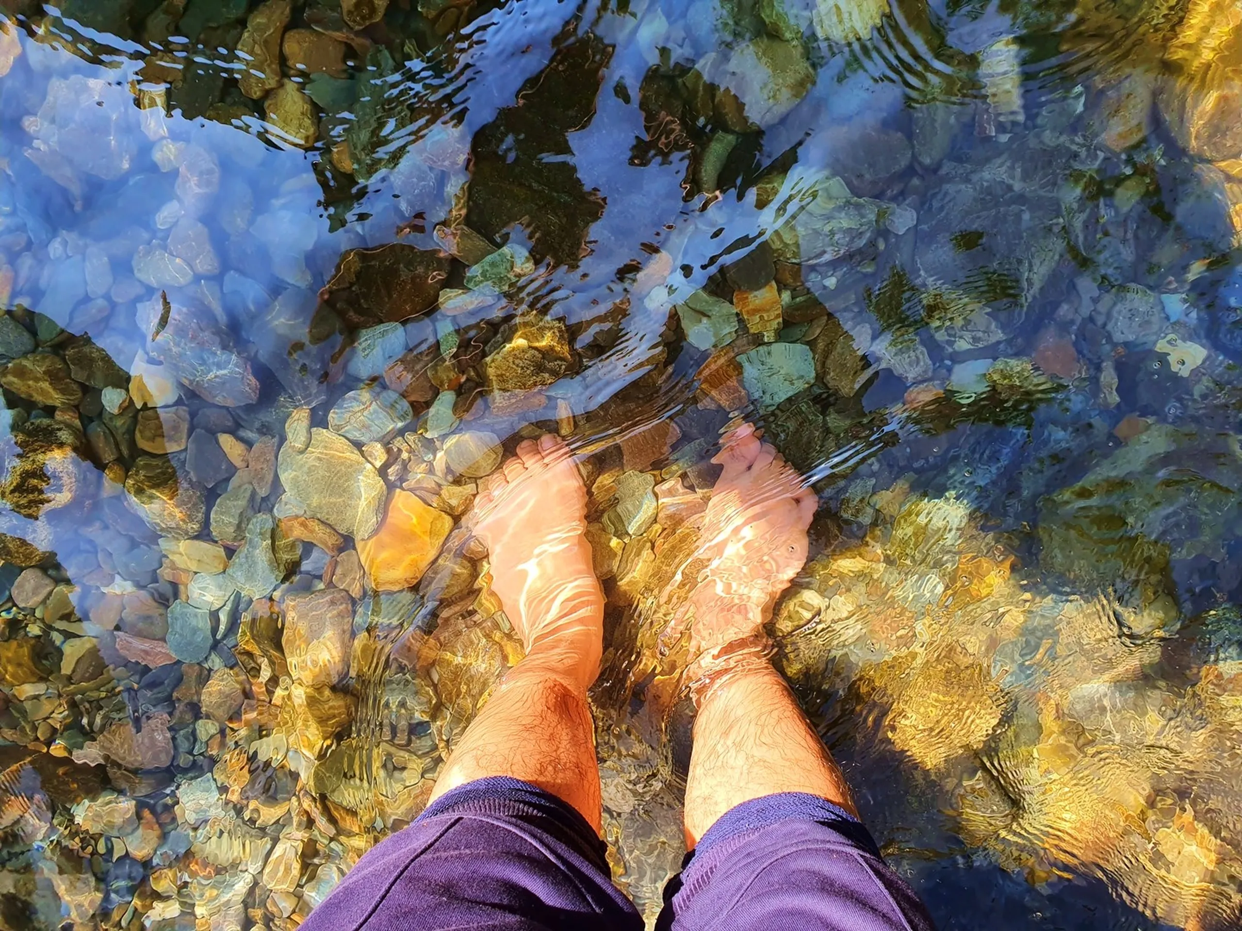 A first-person overhead perspective of bare feet submerged in a crystal-clear mountain stream in Nepal, the colorful pebbled riverbed visible through the shallow transparent water casting sun-ripple reflections of blue, gold, amber, and green - a meditative and refreshing moment of soaking tired trekking feet in a natural Himalayan stream on the Annapurna Circuit route, Nepal - perfect for mindfulness travel, nature photography, and Nepal trekking lifestyle content