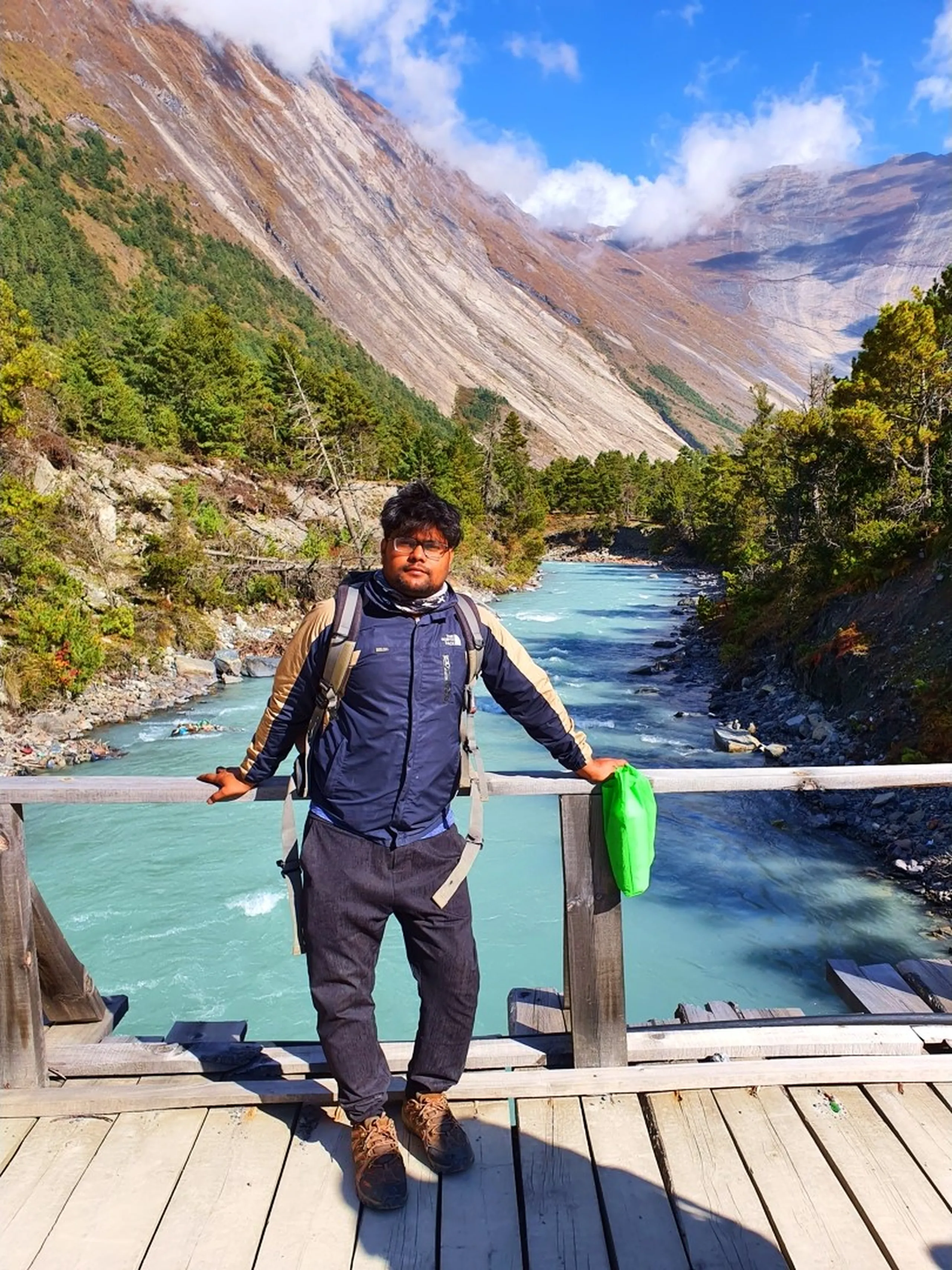 A young male Nepali trekker stands on a wide wooden footbridge over the breathtakingly turquoise Marsyangdi River in Manang, Nepal - wearing glasses, a navy blue and tan North Face two-tone trekking jacket, dark trousers, brown hiking boots, and a loaded backpack with a green bag hanging from it - arms spread wide on the wooden railing in a relaxed, open pose - the luminous glacier-fed river stretches away in both directions in vivid turquoise-blue - pine trees with autumn foliage line both banks - a towering grey and rust Himalayan valley wall rises steeply in the background under a blue sky with white cloud - a stunning Nepal trekking river bridge portrait in the Manang valley, Annapurna Circuit, capturing the sheer natural beauty of the glacial rivers and mountain scenery on the way to Ice Lake Nepal