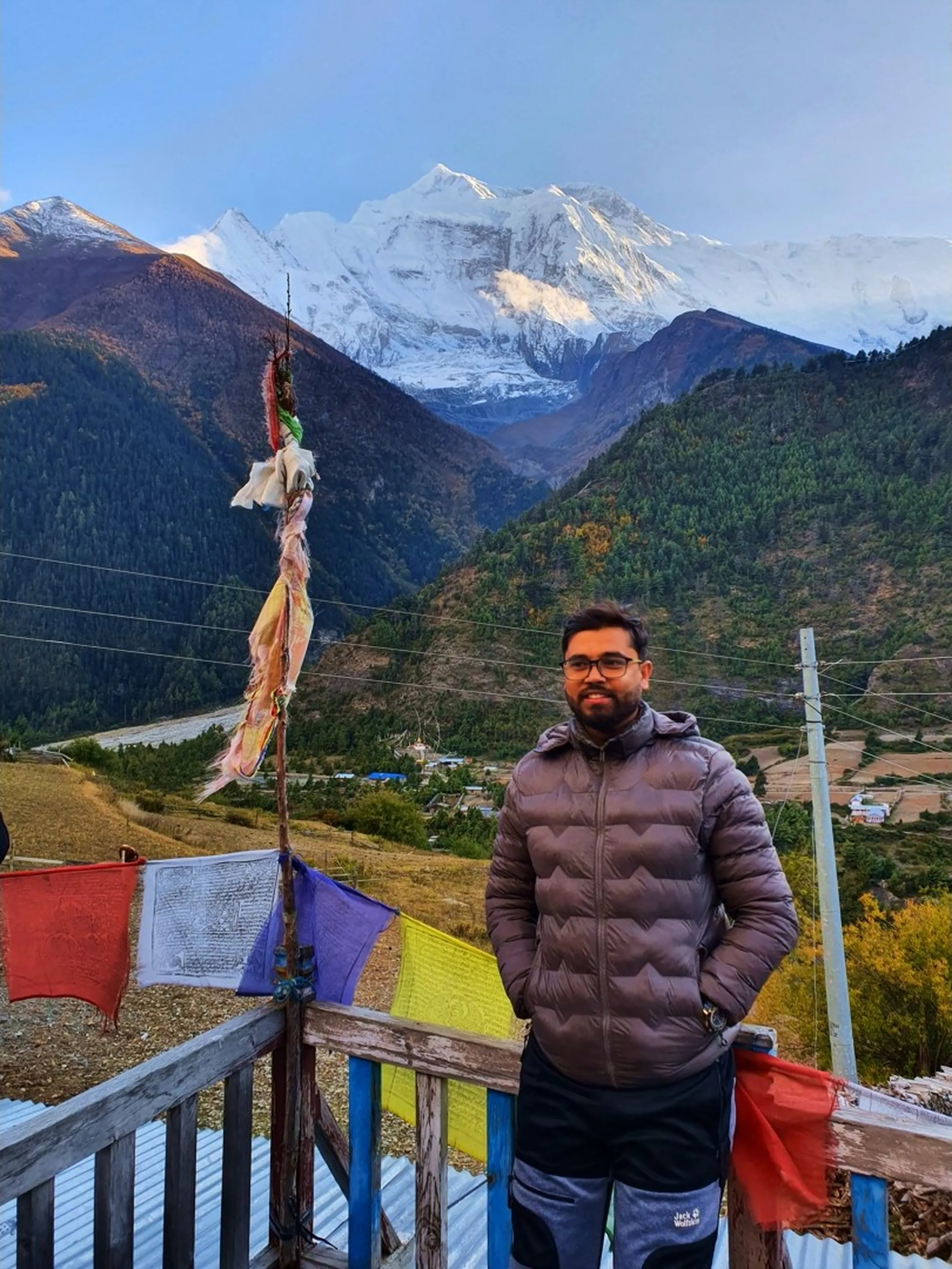 Trek coordinator standing on a wooden terrace in a puffer jacket in front of colorful Tibetan Buddhist prayer flags, with a dramatic snow-covered Annapurna II mountain massif dominating the horizon, pine forest valley and Lower Pisang village scattered below at sunrise - Annapurna Circuit Nepal guide, Upper Pisang viewpoint, Himalayan sunrise prayer flags