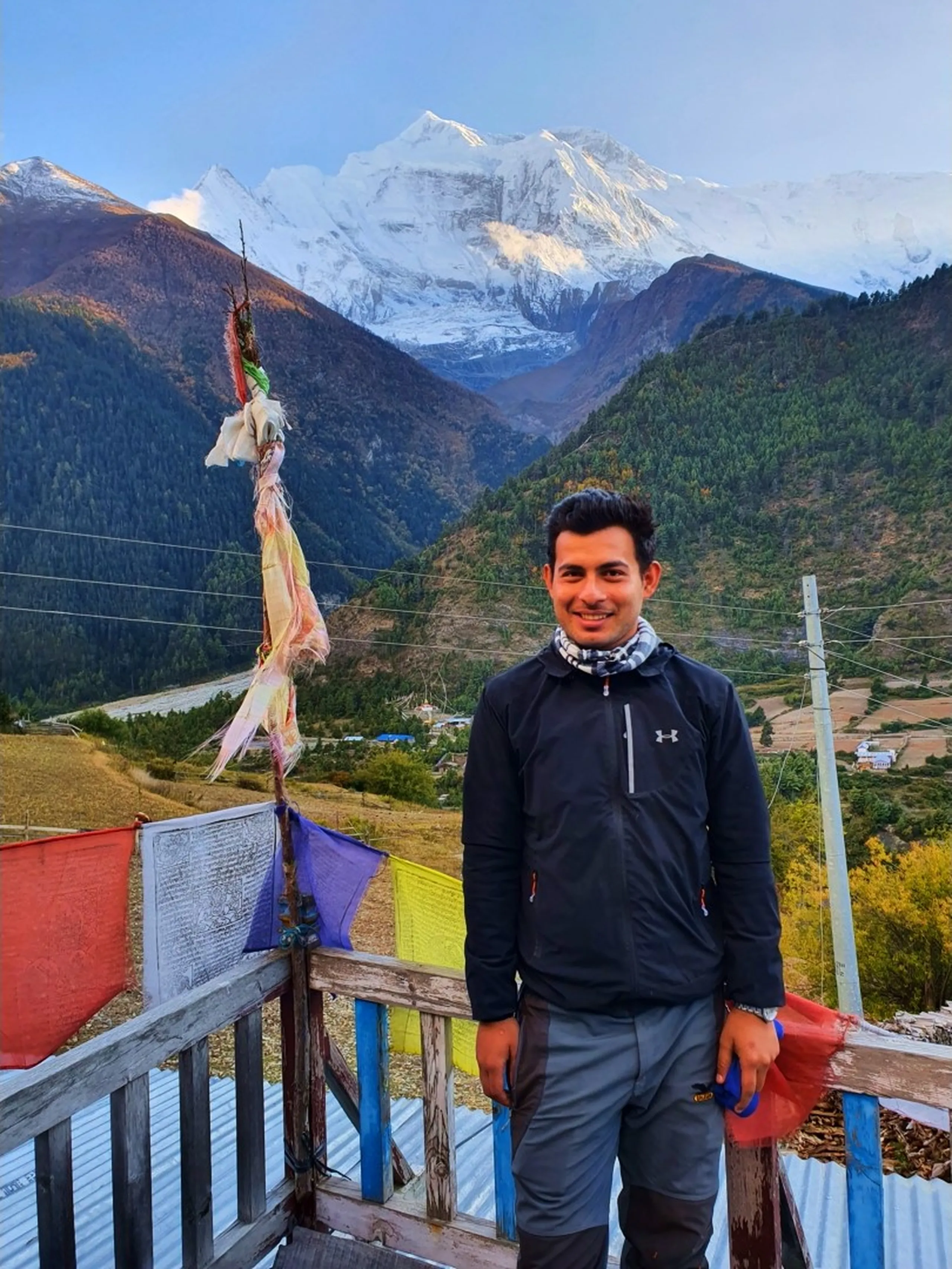 Smiling male trekker in a black Under Armour fleece jacket and scarf posing on a wooden deck with colorful prayer flags at Upper Pisang, Nepal, massive glaciated Annapurna peak gleaming in the background against a pre-dawn blue sky, valley and terraced fields below - Annapurna trekking Nepal, Upper Pisang sunrise, Himalayan adventure travel
