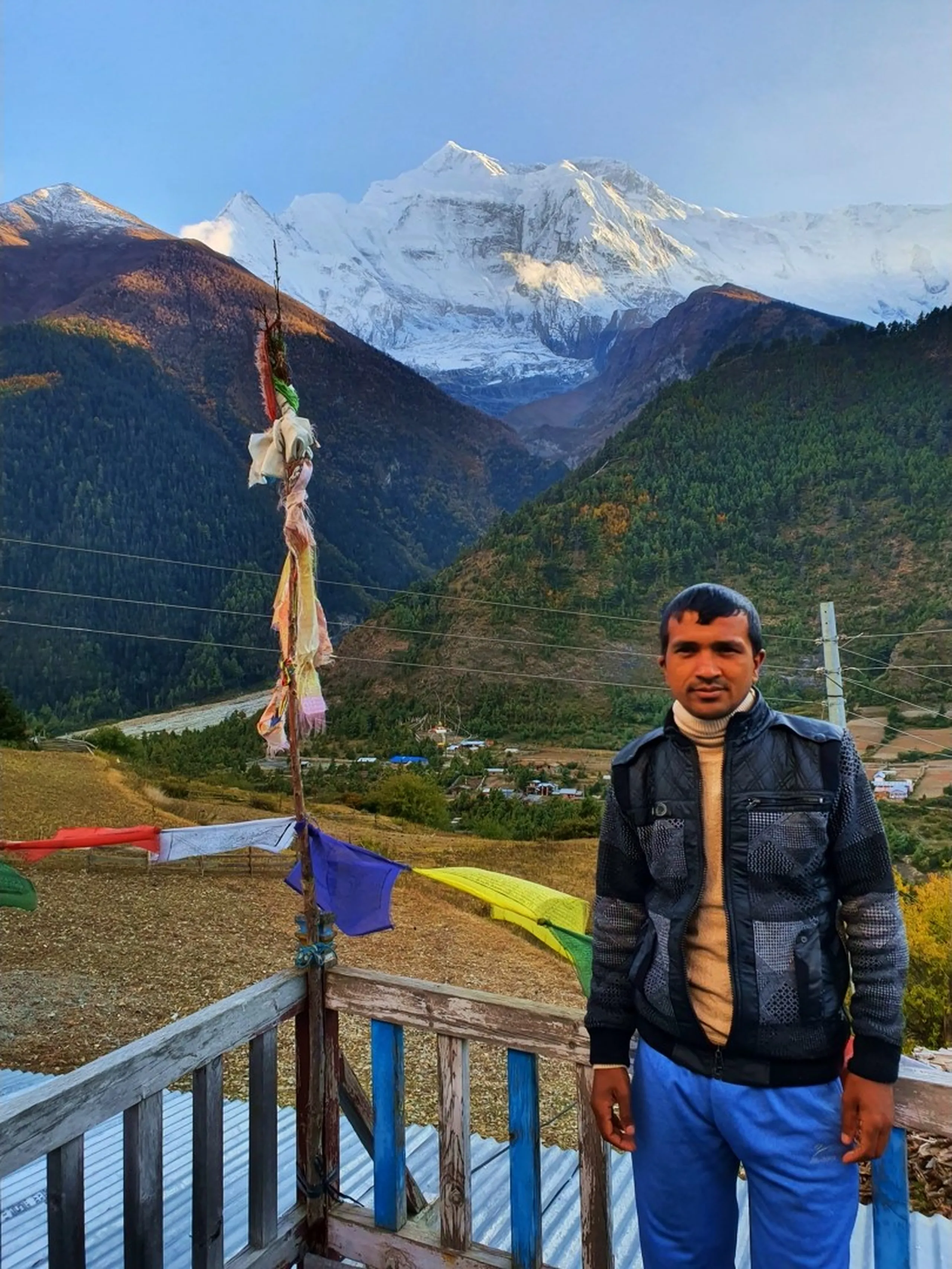 Serious-looking male trek companion in a black quilted vest over a cream turtleneck sweater standing in front of prayer flag poles at Upper Pisang, Nepal, with the vast snow-covered Annapurna range filling the sky behind him and a wide Himalayan valley at dusk - Nepal high altitude trekking, Manang region mountain views, Annapurna Circuit companions