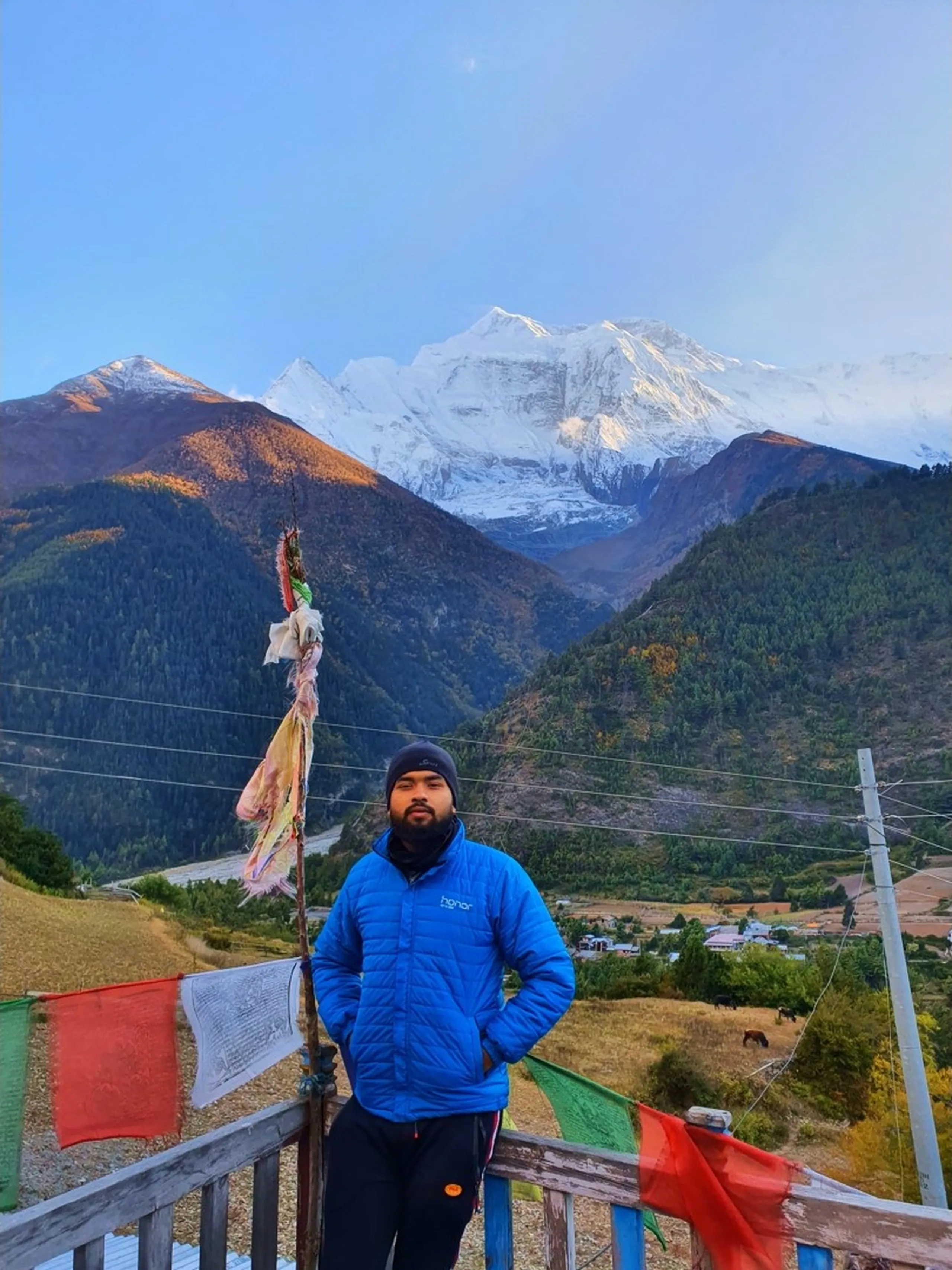 Male trekker in a bright blue Honor-branded puffer jacket and black beanie standing with hands in pockets on a wooden railing terrace at Upper Pisang, Nepal, with red and white prayer flags fluttering, snow-dusted Annapurna peaks glowing in soft alpine light, a grazing horse visible in the valley far below - Annapurna Circuit trek Nepal, Upper Pisang altitude 3310m, Himalayan trekking group