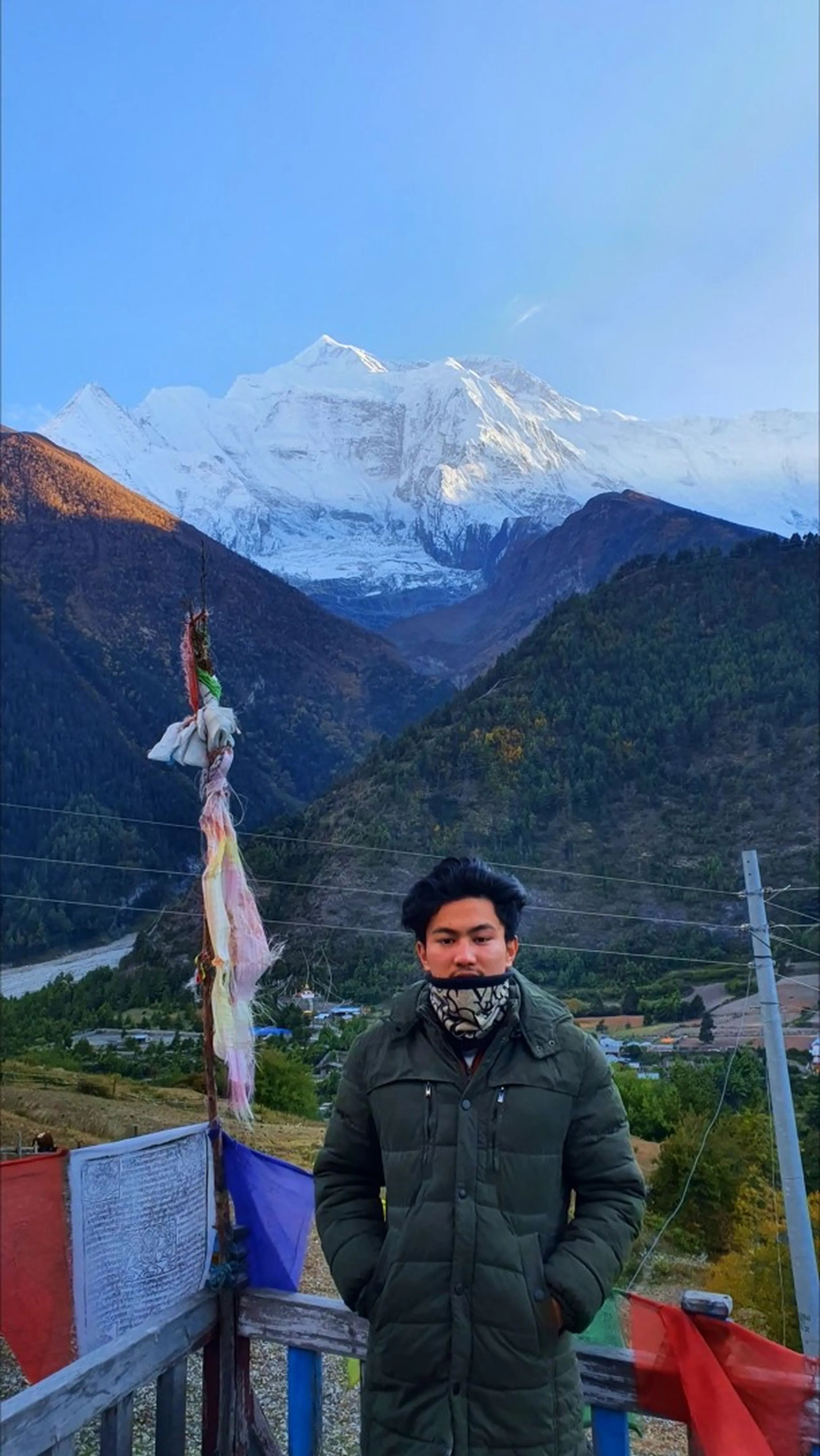Young male trekker in a dark olive green long puffer coat and patterned neck gaiter standing solemnly on a viewpoint terrace at Upper Pisang Nepal, large Buddhist prayer flags draped below, the towering white Annapurna snowfield and glaciers perfectly centred behind him in early morning blue-hour light - Upper Pisang Nepal sunrise, Annapurna II viewpoint, Himalayan trek portrait