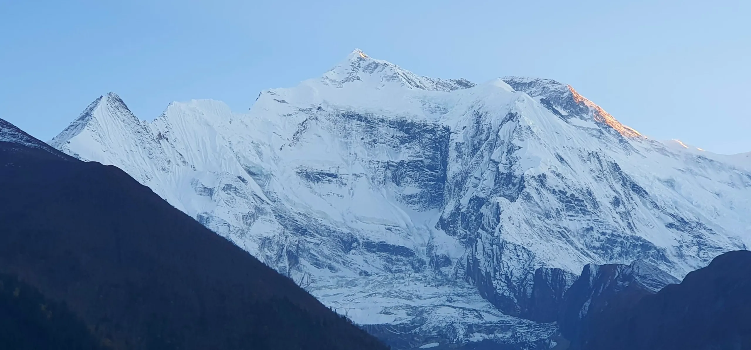 Close-up telephoto shot of the Annapurna II snowface at dawn, enormous white glaciated ridgelines and couloirs bathed in the first warm orange alpenglow on the right flank, with smaller rocky satellite peaks to the left against a pale powder-blue pre-sunrise sky - Annapurna II Nepal photography, Himalayan glacier close up, best mountain photography Nepal