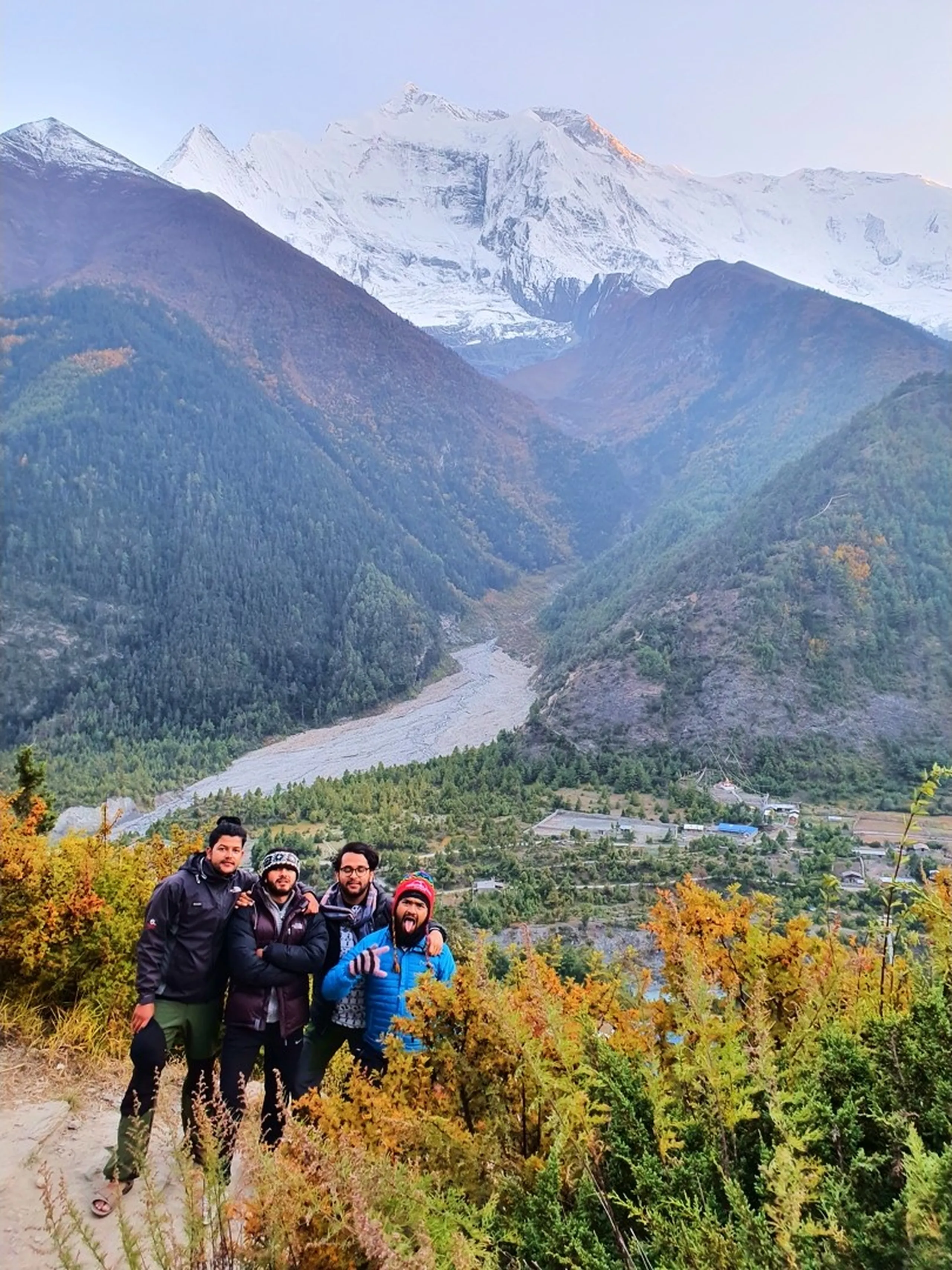 Group of four male trekkers posing playfully on an uphill dirt trail above Upper Pisang village, Nepal, with one sticking out his tongue, all bundled in trekking jackets; behind them a wide river valley fringed with autumn orange and gold foliage leads the eye toward the towering snow-capped Annapurna massif glowing pink at first light - Annapurna Circuit group trek, friends trekking Nepal, Upper Pisang to Manang trail