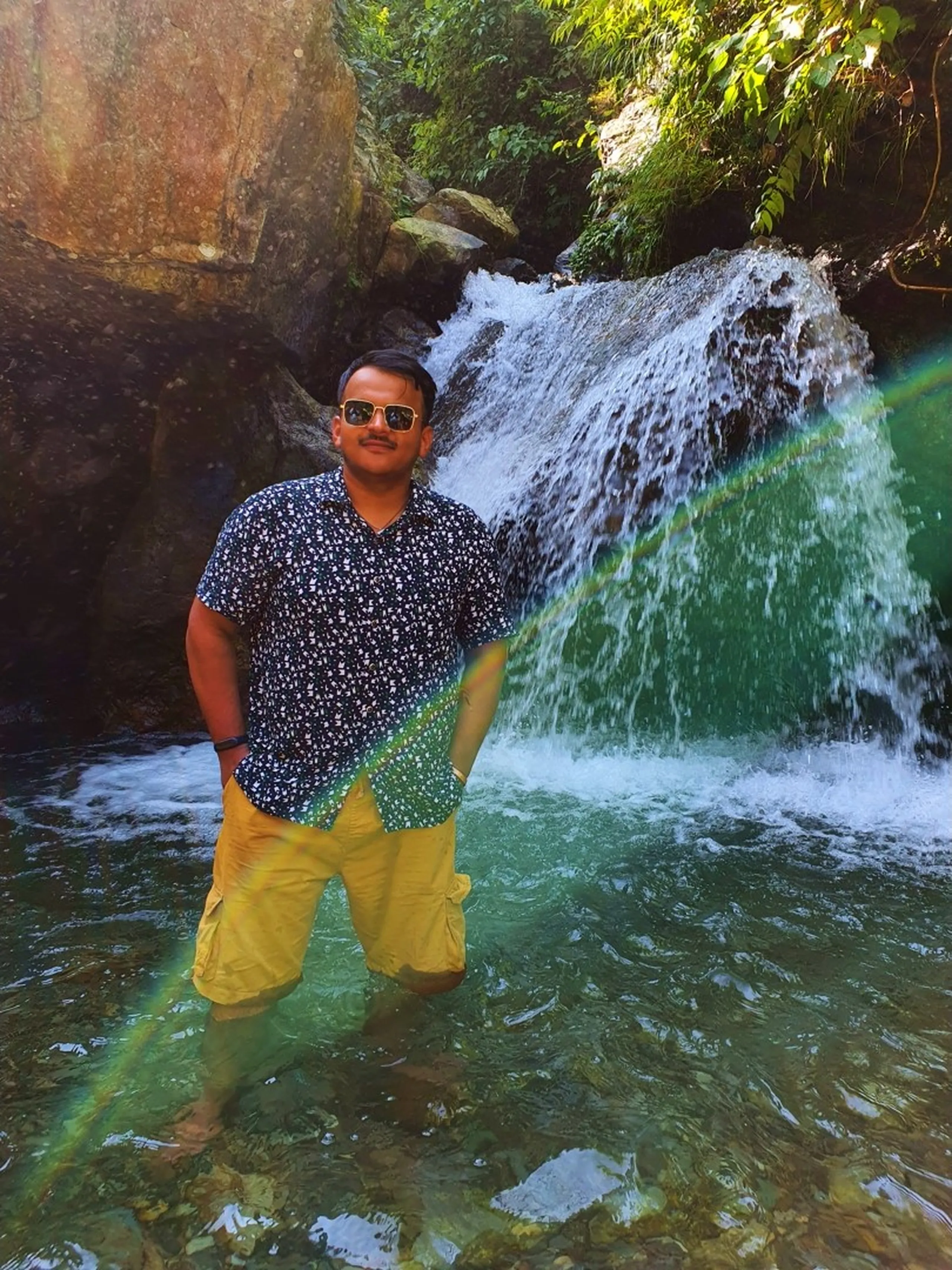 A young male trekker stands knee-deep in the turquoise pool of a cascading waterfall in Nepal, wearing a patterned navy short-sleeve shirt, yellow cargo shorts, and aviator sunglasses, hands in pockets and smiling confidently - white rushing water spills over mossy dark boulders behind him, sunlight creating a natural rainbow lens flare across the frame - lush jungle greenery framing the top - a stunning Nepal waterfall portrait on the trek to Lunjung, Lamjung, ideal for Nepal hidden waterfall and Annapurna trekking blog content