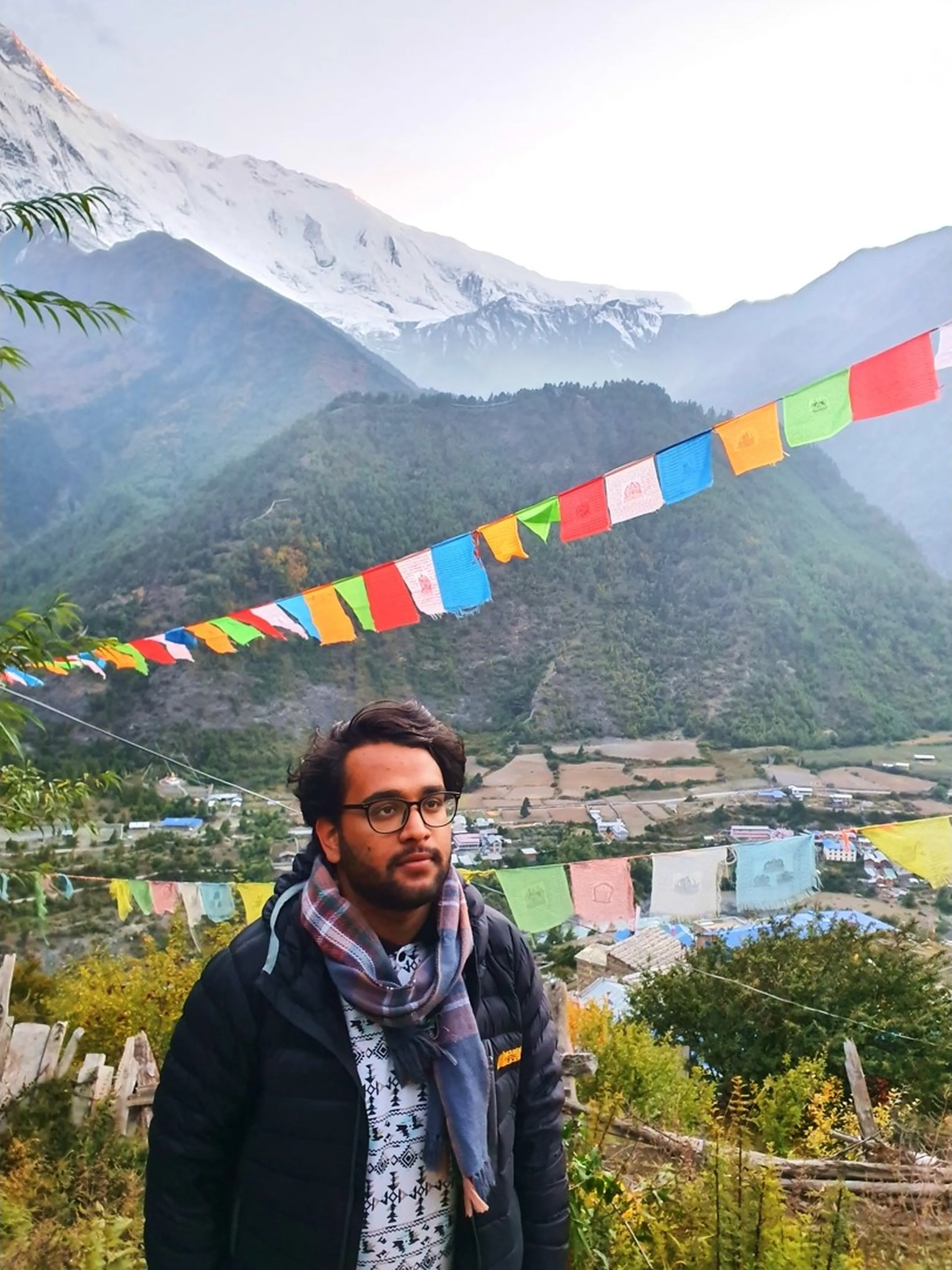 Solo male trekker with glasses and a plaid scarf looking contemplatively to the side, standing beneath a line of vibrant multicolour Tibetan prayer flags strung across a hillside viewpoint with Lower Pisang village and terraced farmland spread out below, misty Annapurna peaks partially veiled in cloud behind - Nepal prayer flags Annapurna, Manang valley trekking, solo Himalayan trekker