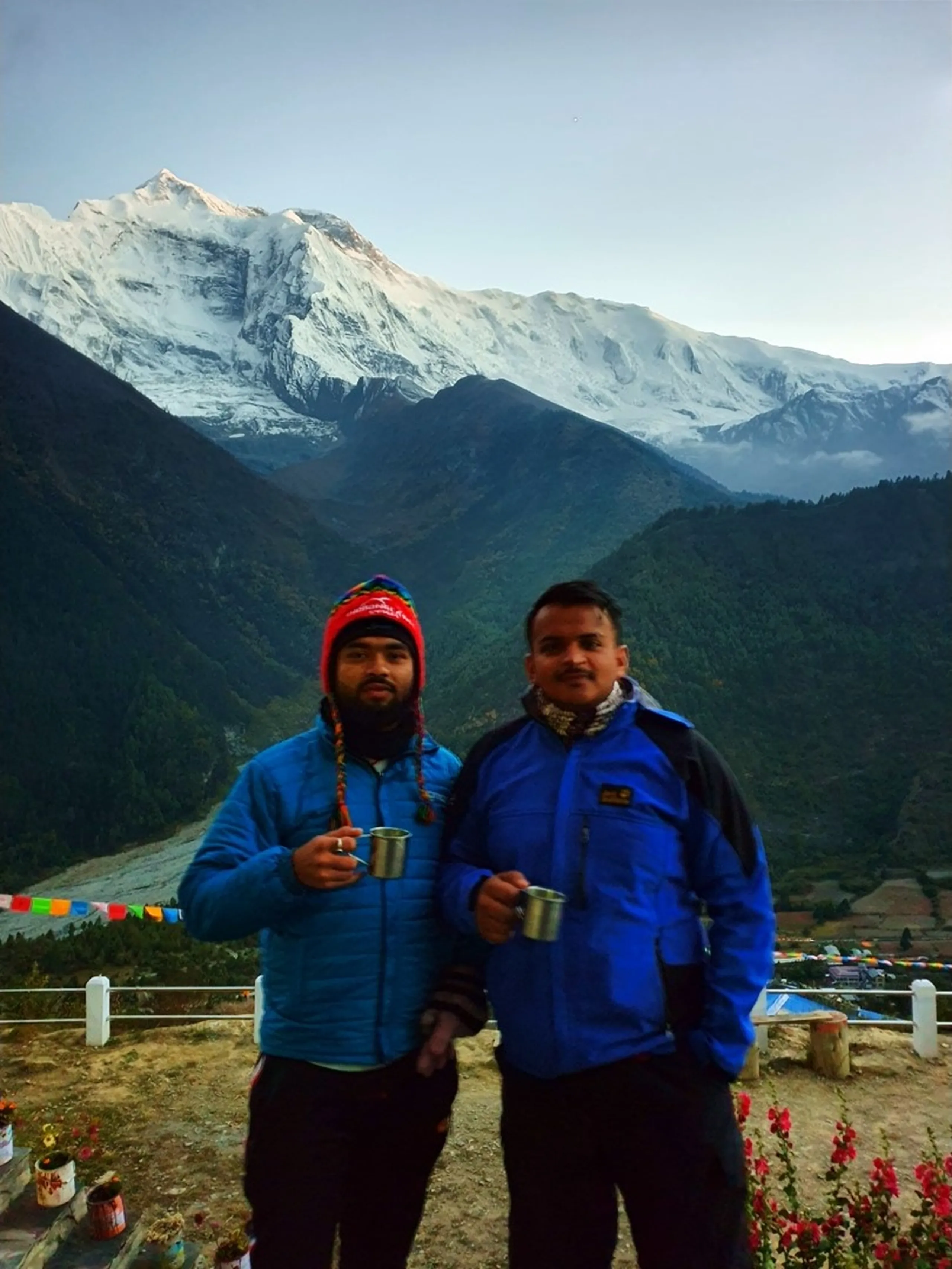 Two male trekkers holding metal cups of hot tea or local brew on a rooftop viewpoint in Manang Nepal, one wearing a colourful braided Chullo hat and blue puffer jacket, the other in a blue Jack Wolfskin fleece, both smiling warmly; the spectacular Annapurna massif stretches across the entire background above the Marsyangdi river valley with red flowers in the foreground - Annapurna Circuit Manang tea break, trekking friends Nepal, high altitude refreshment with mountain view