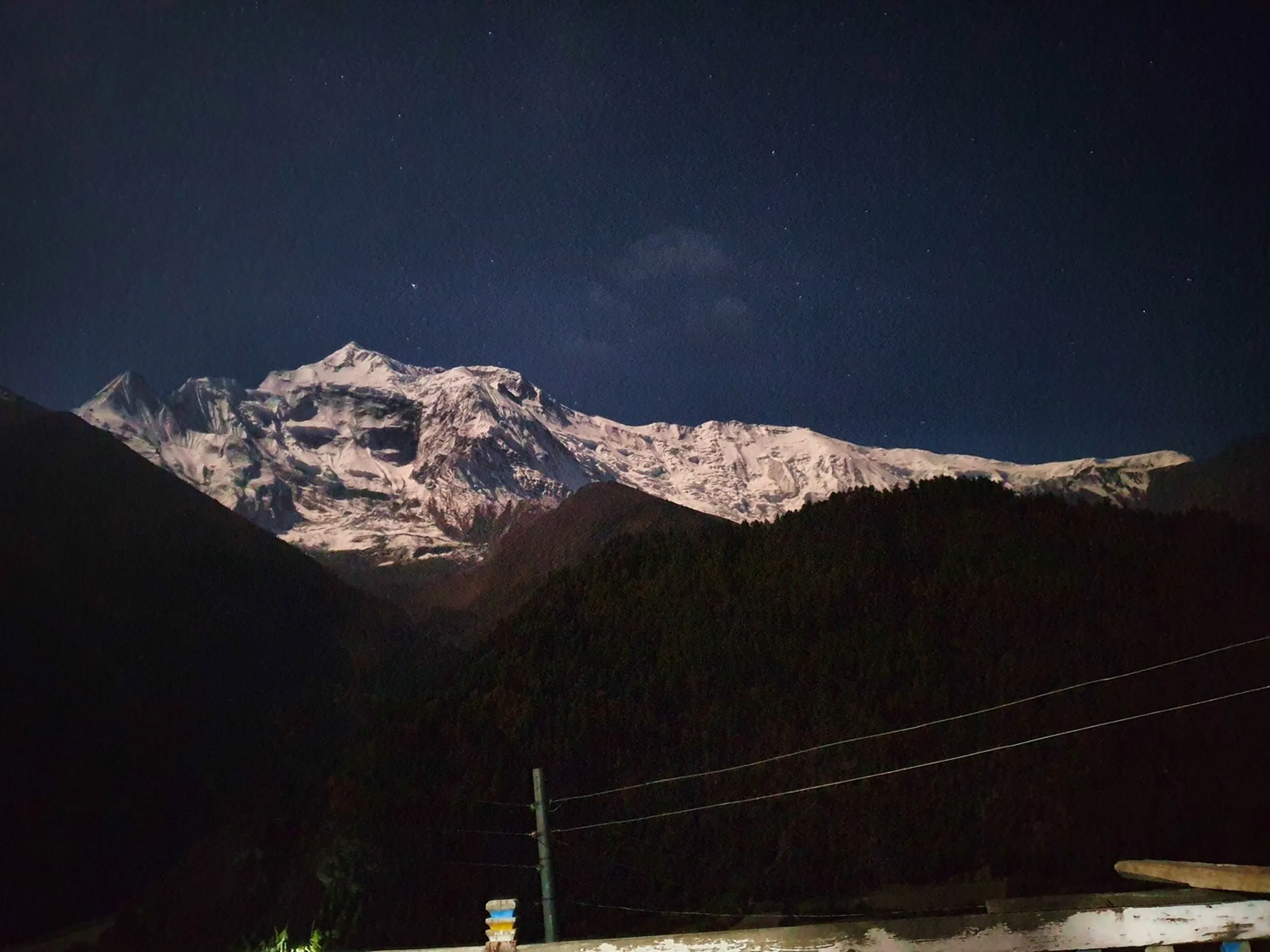 Dramatic Himalayan night photography of Annapurna snowfields glowing under natural moonlight against a deep navy star-speckled sky, dark pine forest silhouette in the foreground, electric power lines crossing the frame - Annapurna night photography Nepal, stargazing Himalaya, Manang astrophotography