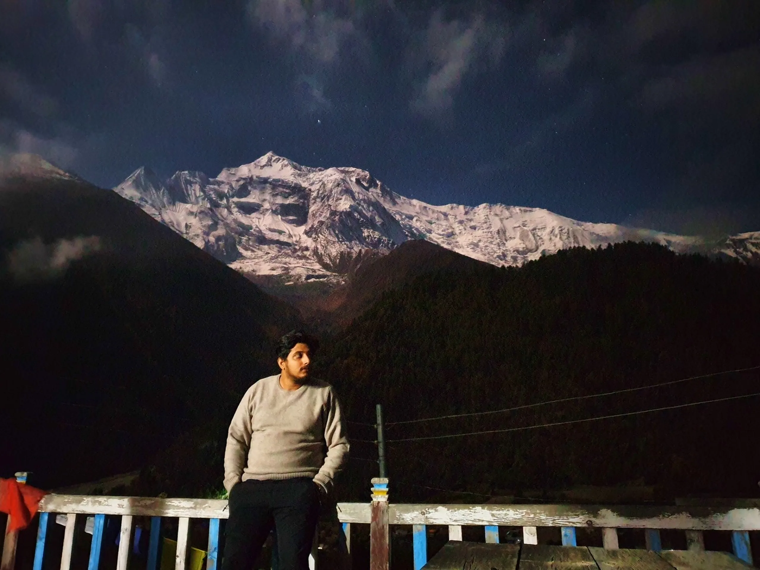 Male trekker in a grey sweater sitting on a blue and white wooden railing at night in Upper Pisang Nepal, looking away pensively, the Annapurna glacier face luminously lit by moonlight above dark forest silhouettes under a partially cloudy star-filled sky - Himalayan night trek portrait, Upper Pisang Nepal nightscape, moody mountain photography