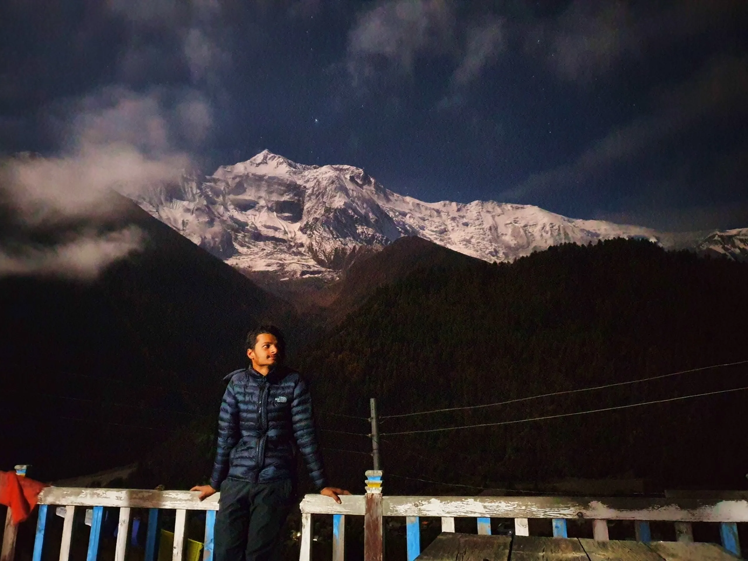 Young male trekker in a navy blue North Face down jacket leaning on a weathered white railing at night with moonlit Annapurna snowfields rising dramatically through clouds behind him, a single star visible in the deep blue sky - Nepal night mountain view, Annapurna moonlit peak, high altitude trekking night photography