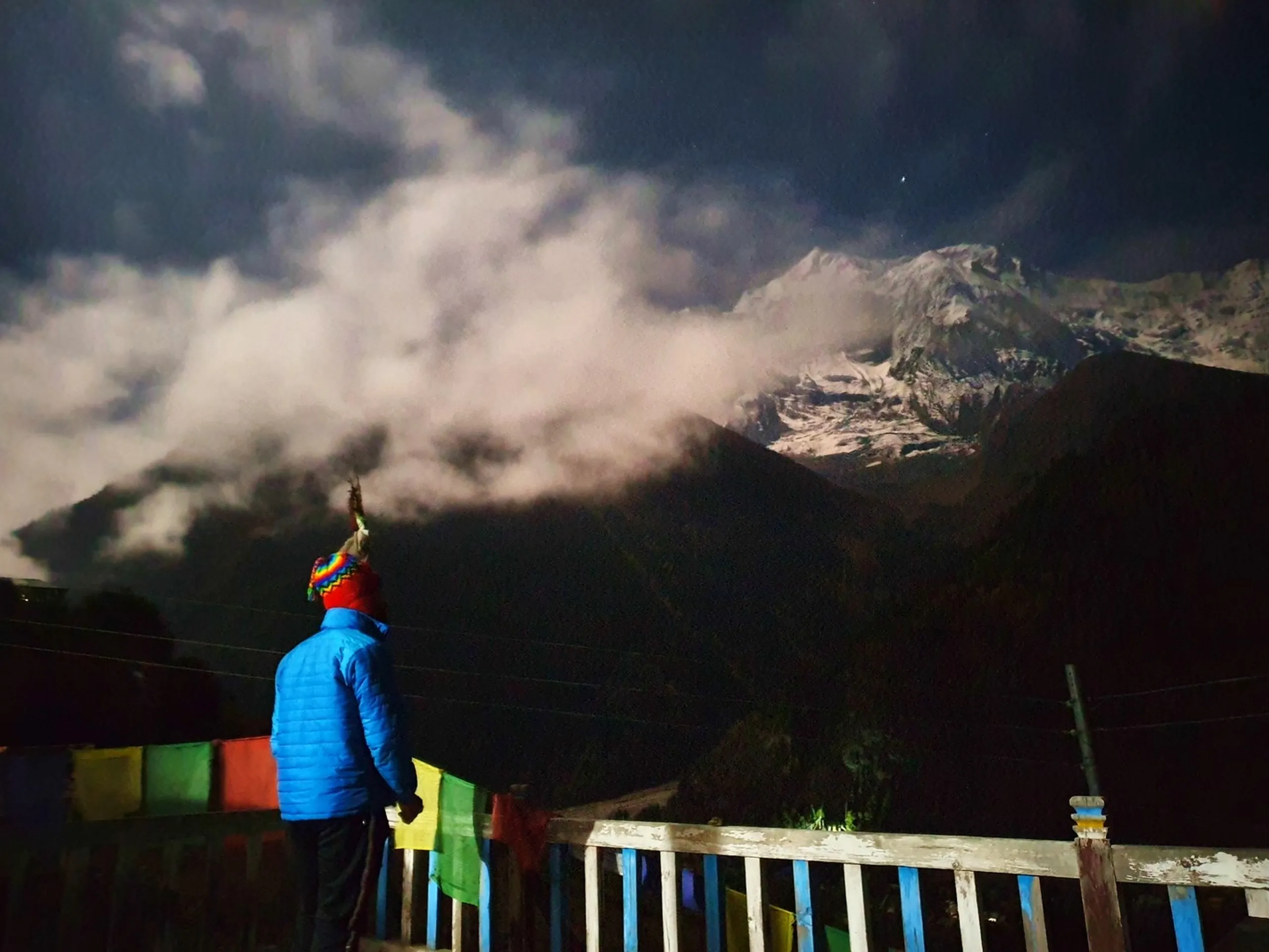 Back view of a trekker in a bright blue down jacket and colourful Bolivian-style Chullo hat standing at a wooden railing terrace gazing up at Annapurna snowfields partially shrouded in dramatic swirling night clouds, colourful prayer flags glowing below in the darkness - Nepal night sky trekking, Annapurna clouds at night, contemplative Himalayan adventure