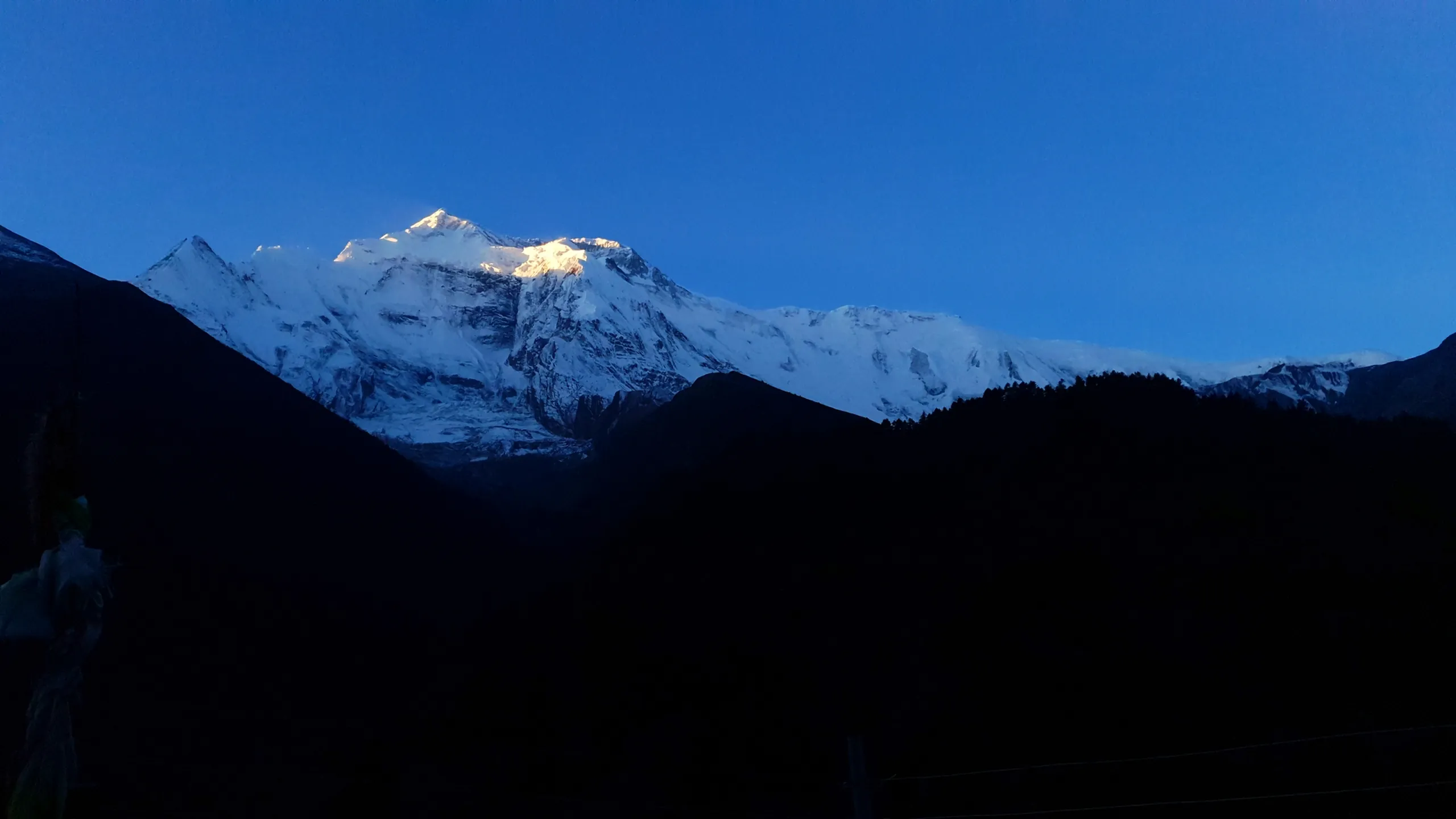 Pre-dawn blue hour panorama of Annapurna II and Annapurna IV from Upper Pisang Nepal, summit ridgeline just catching the first warm golden alpenglow of sunrise while the valley and lower peaks remain in deep blue shadow, prayer flags barely visible at left edge - Annapurna sunrise blue hour, best Nepal mountain dawn photography, Annapurna Circuit early morning