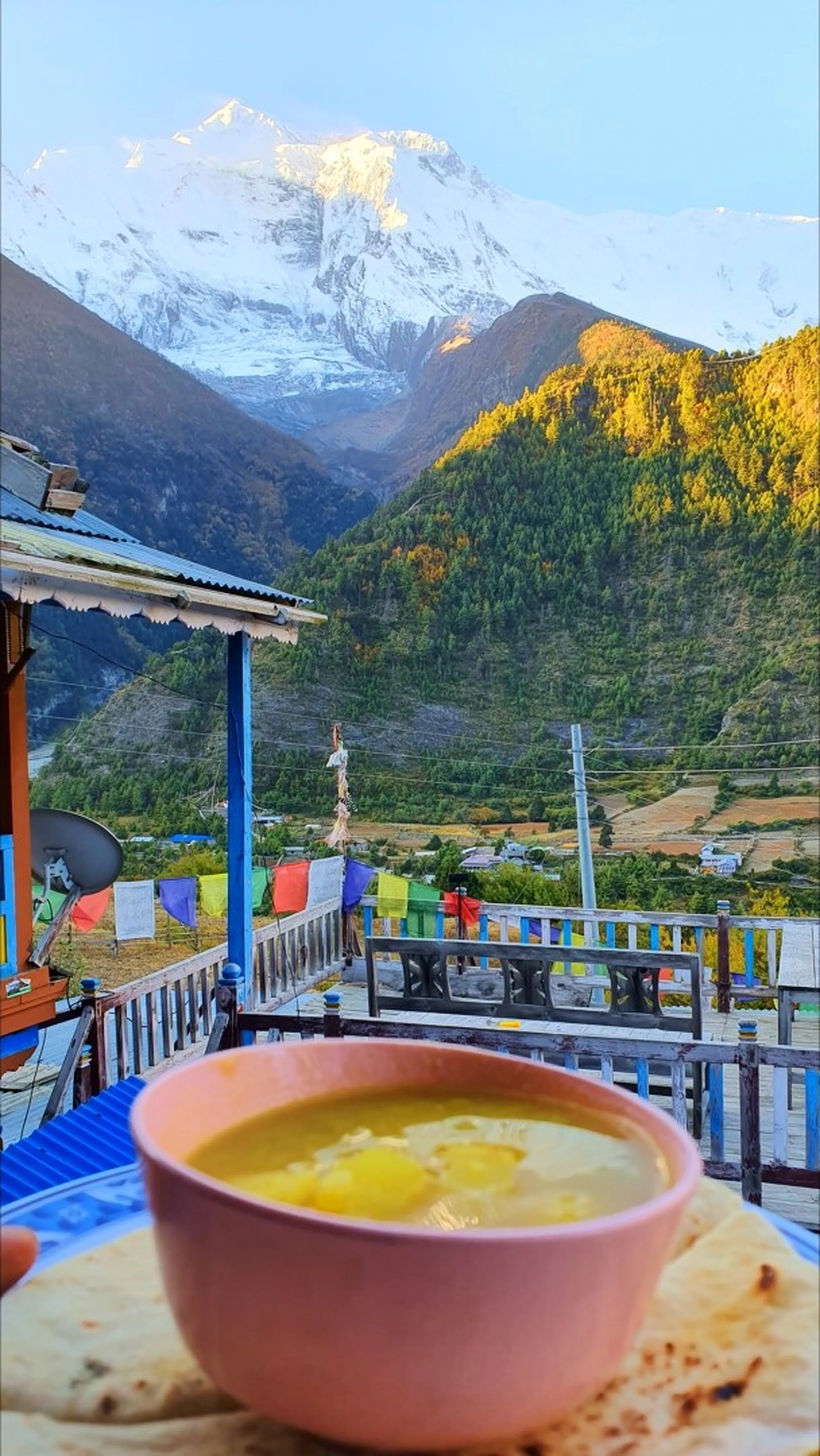 Last breakfast at a guesthouse terrace in Upper Pisang Nepal before departing for Manang, a bowl of warm potato or lentil soup with flatbread in the foreground, the Annapurna range spectacularly lit in warm morning alpenglow behind the Marsyangdi valley, colourful prayer flags strung across the terrace - Nepal trek food mountain view, Himalayan guesthouse breakfast, Annapurna Circuit dal bhat