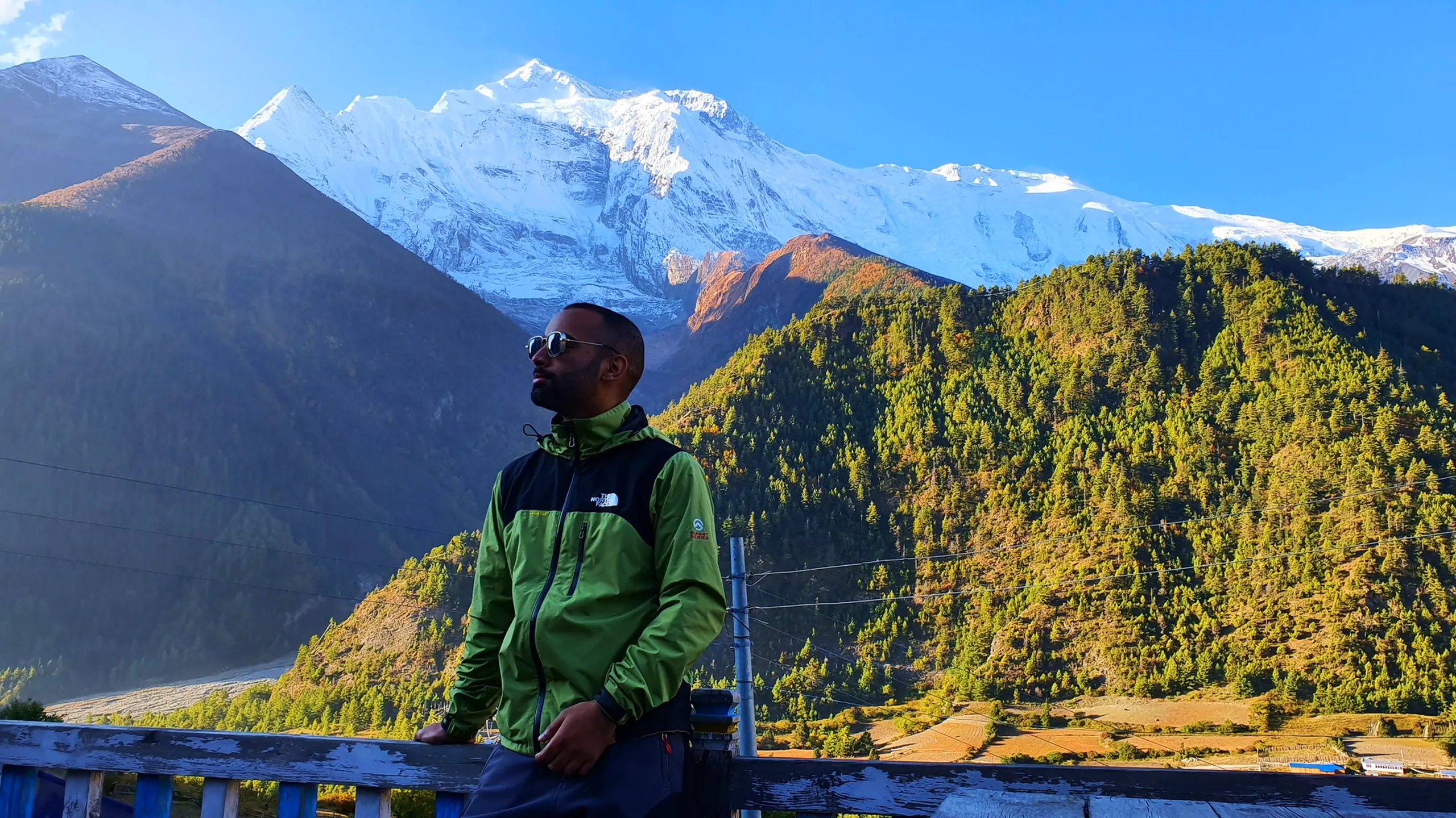 Male trekker in a green and black North Face softshell jacket wearing mirrored silver sunglasses, leaning against a wooden railing and looking to the side in a relaxed pose, the wide Annapurna range blazing white against clear blue sky with autumn-tinted pine-covered hills below in warm morning light - Nepal trekking style, Annapurna Circuit Manang viewpoint, Himalayan adventure portrait