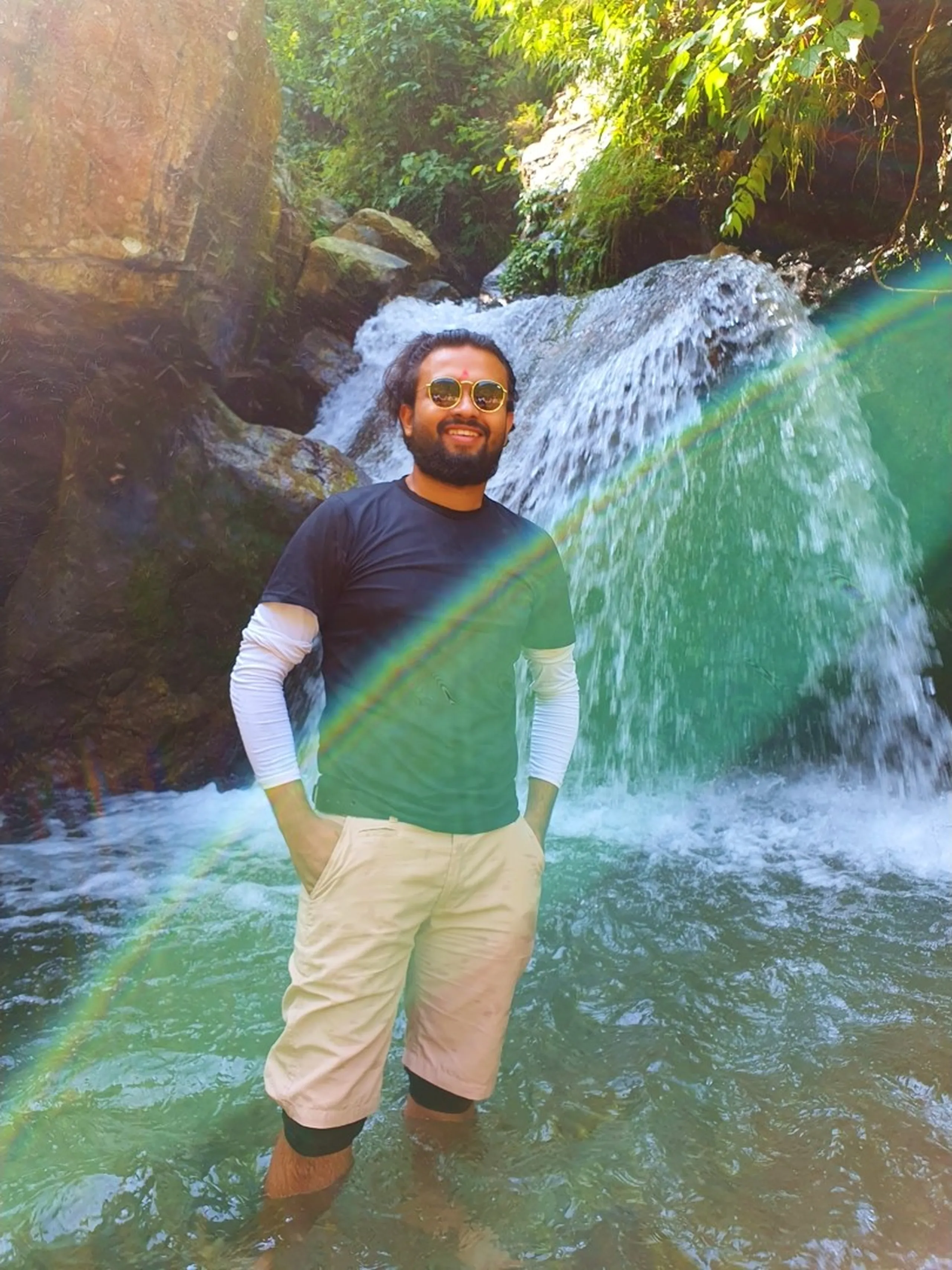 A young male trekker stands smiling in the shallow turquoise pool at the base of a Nepal mountain waterfall, wearing a dark navy t-shirt layered over a white long-sleeve, beige shorts rolled above the knees, and yellow-framed aviator sunglasses, with a red tika on his forehead - a dramatic rainbow lens flare cuts diagonally across the frame as white water cascades over dark boulders behind him - bright jungle foliage overhead - a vibrant and colorful Nepal waterfall photography moment on the Lunjung trek, Annapurna region