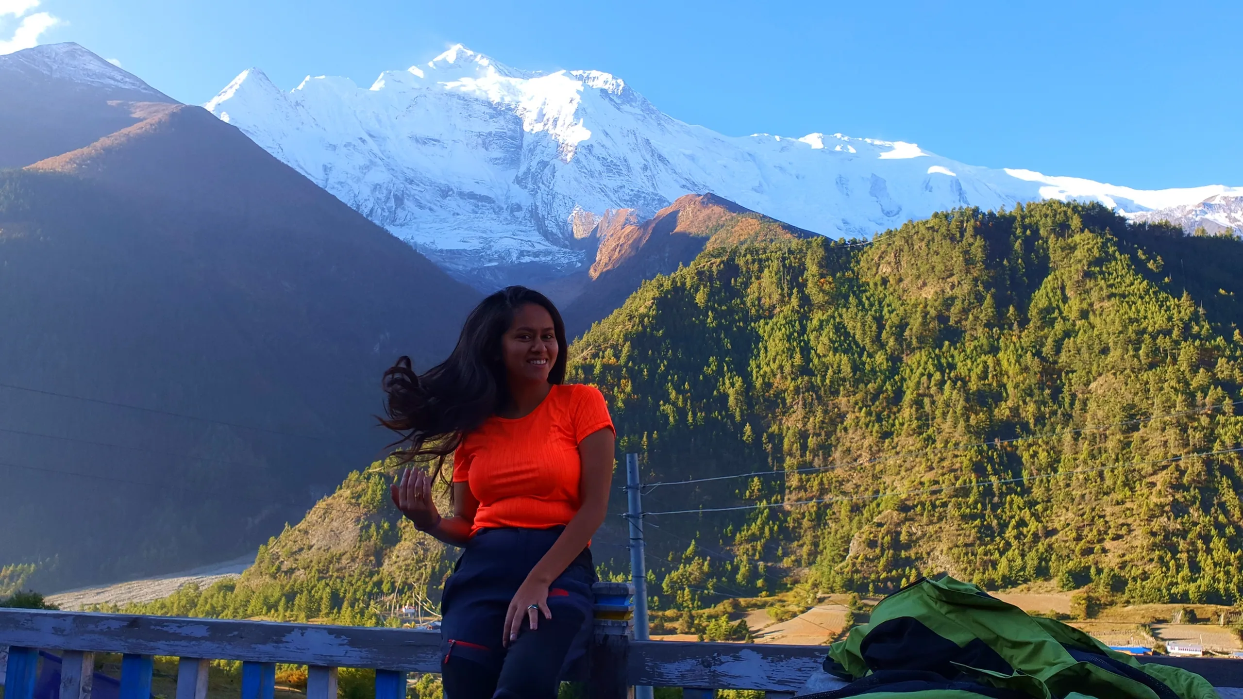 Joyful female trekker in a vibrant orange fitted top sitting on a blue wooden railing with her long dark hair caught mid-toss by the mountain wind, smiling freely; the full breadth of the Annapurna snowfields spread across a brilliant blue sky behind her, autumn-coloured pine forests on the valley slopes - woman trekking Annapurna Circuit Nepal, Manang female trekker, best Himalayan trek experience