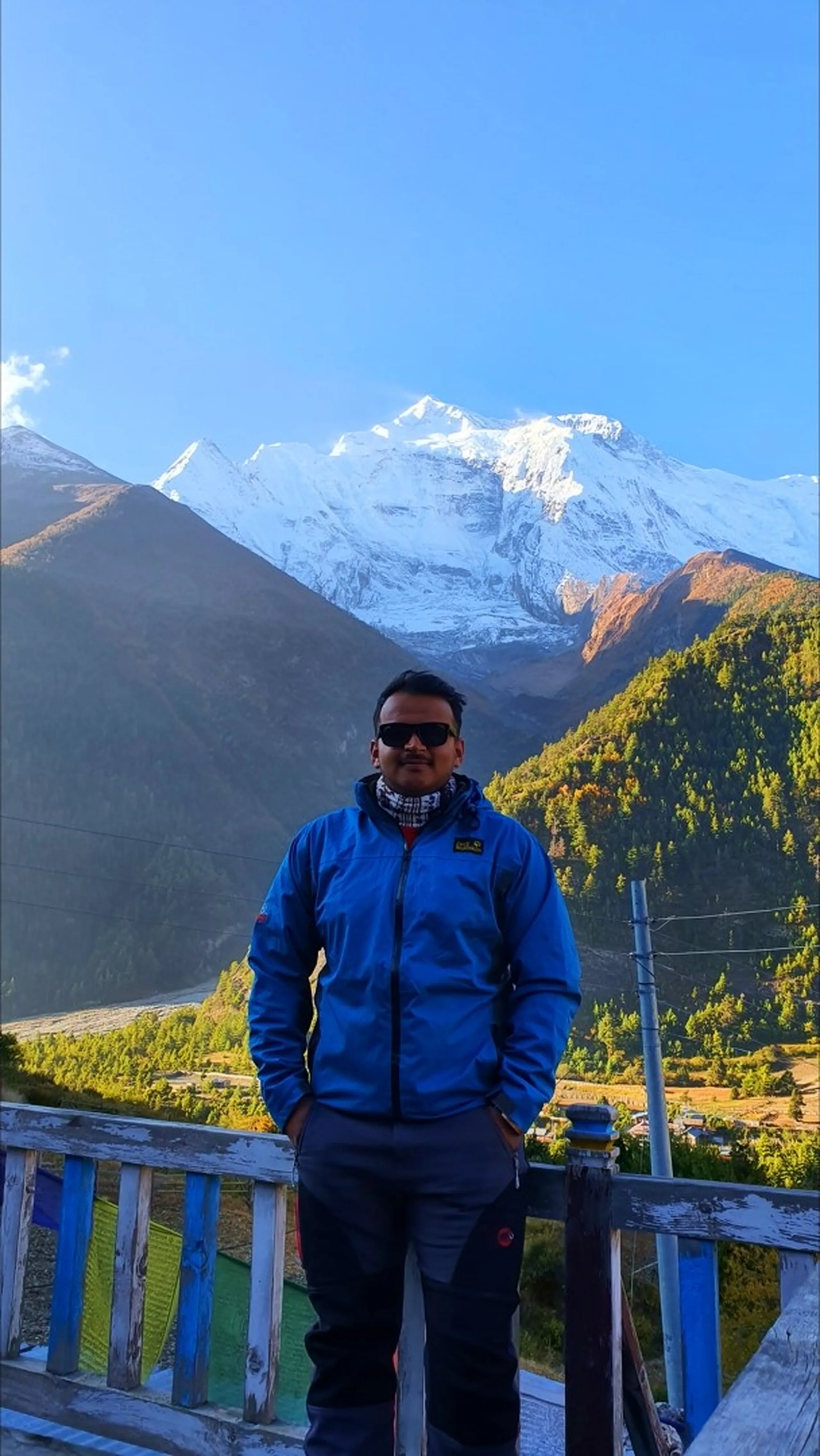 Male trekker in a blue Jack Wolfskin softshell jacket and dark sunglasses standing with hands in pockets on a weathered wooden railing terrace at Upper Pisang, Nepal, with the full Annapurna II snowface glowing white above autumn-golden pine forests and Lower Pisang valley in warm morning light - Annapurna Circuit trek Nepal, Manang region trekking portrait, Himalayan adventure travel