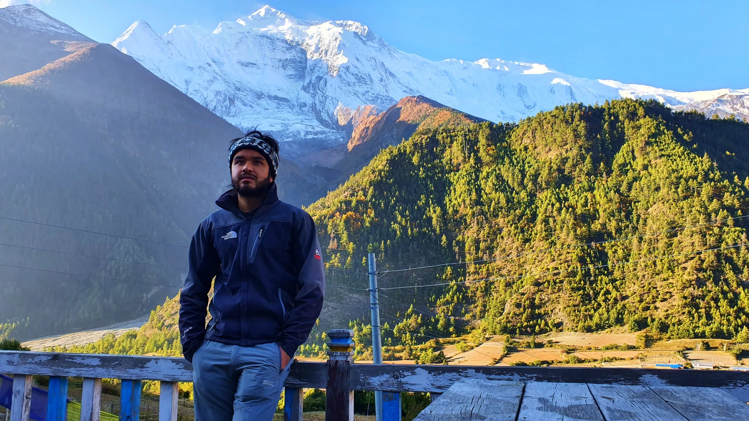 Confident male trekker in a dark navy North Face softshell jacket and patterned headband leaning against a blue wooden railing in Manang Nepal, gazing thoughtfully to the left, the sweeping Annapurna snowfields and autumn pine-covered valley spread magnificently behind him in bright morning sunshine - Annapurna Circuit Manang Nepal, North Face trekking gear Himalaya, best Nepal trek viewpoint