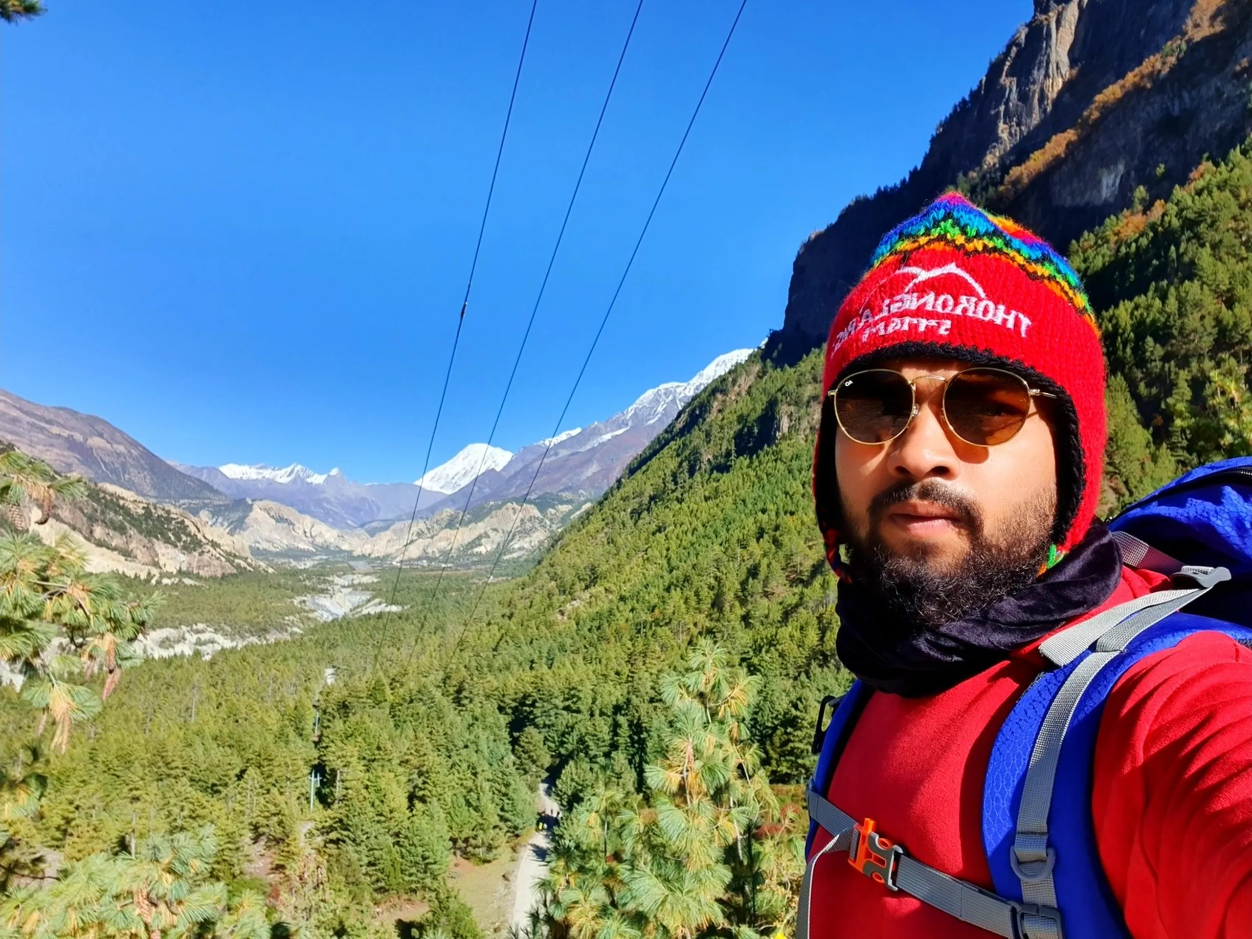Close-up trekking selfie of a bearded male trekker in a red hoodie and colourful Thorong La Pass Chullo beanie hat with rainbow bands and braids, wearing amber aviator sunglasses, blue backpack straps visible on shoulder; wide Marsyangdi river valley with dense pine forests and snow-capped Annapurna peaks receding into the distance behind him under clear blue sky - Thorong La Pass souvenir hat Nepal, Annapurna Circuit selfie, trekking selfie Manang valley