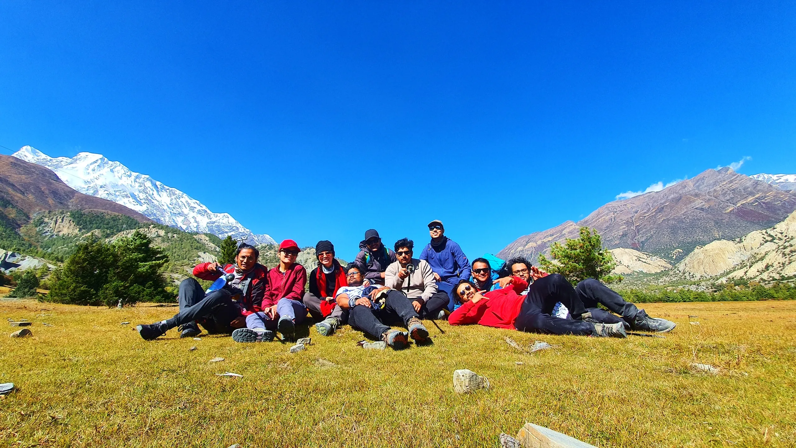 Relaxed group of approximately eleven trekkers lying and sitting together on a sunlit alpine meadow in Manang Nepal for a candid rest stop photo, all smiling and laughing in a casual cluster, trekking poles and backpacks nearby; Annapurna II snowfield visible on the far left, arid cliffs on the right, massive open blue sky filling the upper two-thirds of the frame - Annapurna Circuit group rest stop, trekking friends Nepal, Manang high altitude grassland