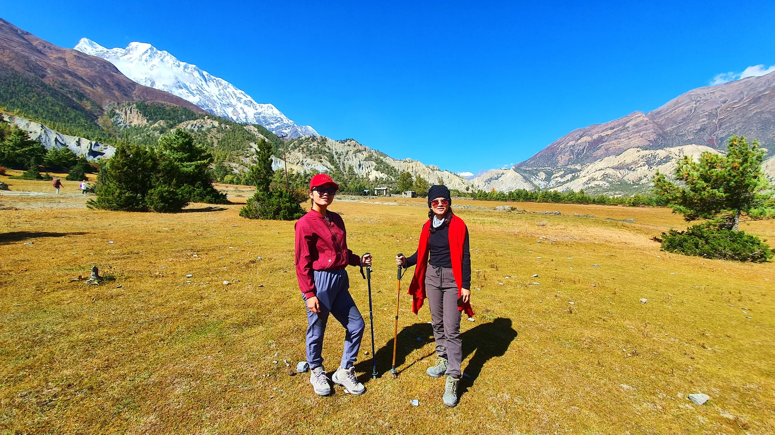 Two female trekkers standing side by side on a golden high-altitude meadow in Manang Nepal, both holding trekking poles - one in a maroon windbreaker and red cap, the other in a black top with a red scarf draped over her shoulders and pink-tinted sunglasses; Annapurna II snowcap gleams behind them on the upper left, eroded Himalayan badlands and pine trees complete the backdrop - women trekking Annapurna Circuit Nepal, female trekkers Manang, Himalayan women adventure
