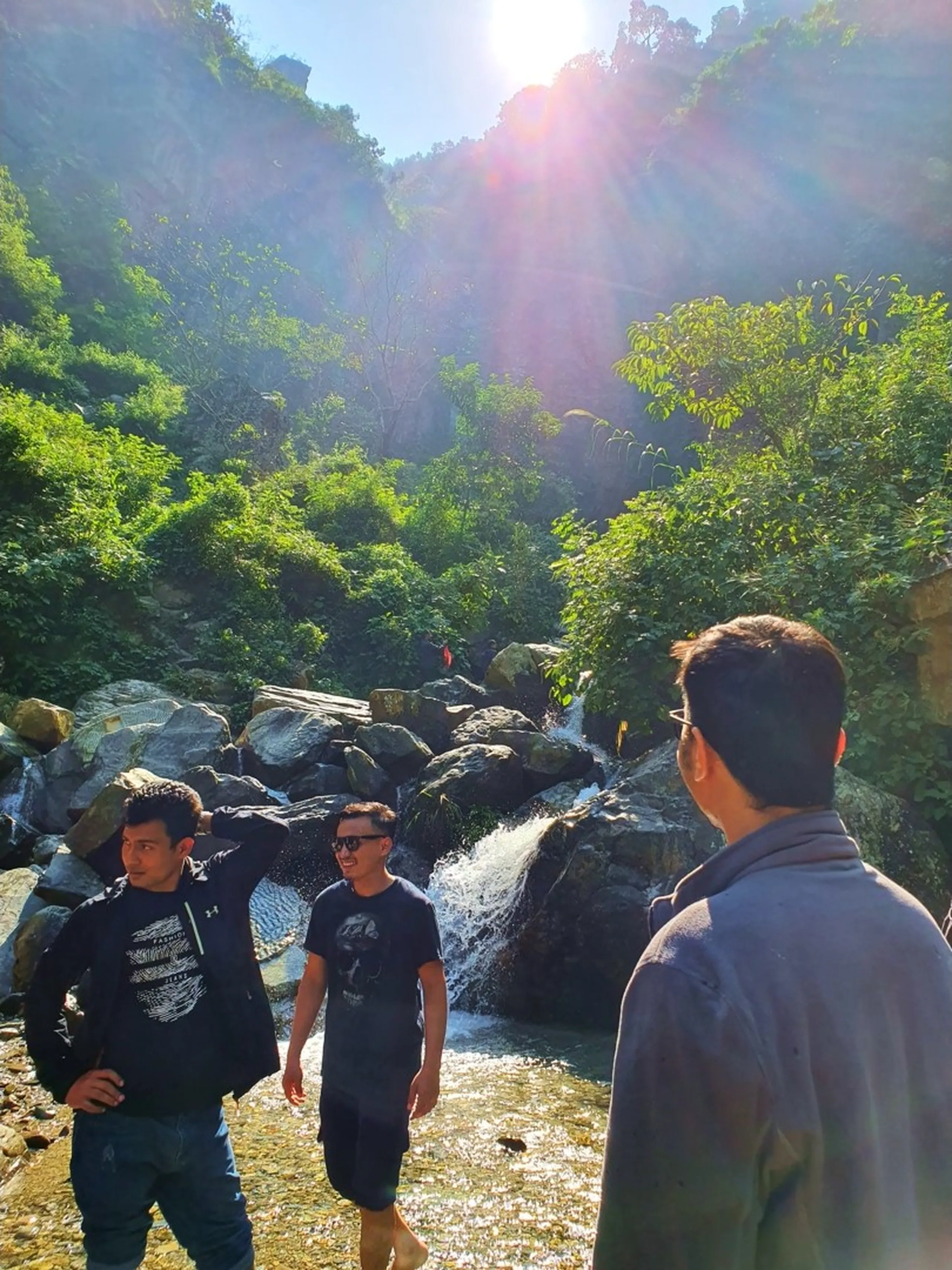 Three male trekkers stand and converse near the rocky banks of a mountain waterfall in Nepal, backlit by dramatic golden sun rays breaking through a narrow gorge of dense green jungle above - white water cascades over large boulders in the background creating misty ambiance - one wearing a black hoodie with sunglasses, another in a black graphic tee barefoot, a third in a grey jacket facing away from camera - a candid and atmospheric Nepal trekking moment with sunburst effect, perfect for Annapurna Circuit trail photography and Nepal adventure travel content
