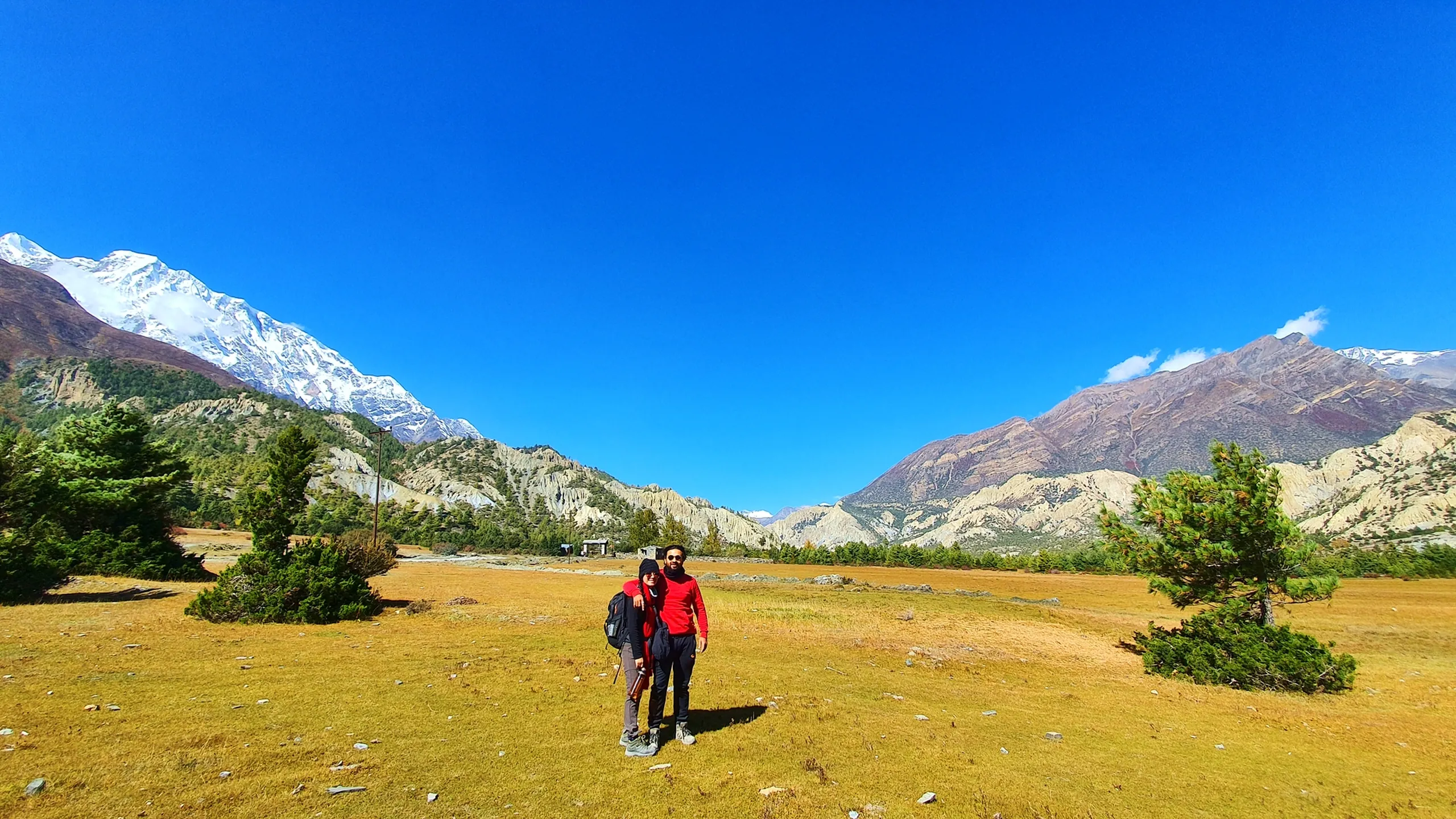 Two trekkers - a woman in a black cap and dark jacket and a man in a red long-sleeve top and sunglasses - standing close together arm-in-arm on a vast golden alpine meadow in Manang Nepal, the Annapurna II snowcap rising on the left and wide arid valley receding behind them, scattered pines on either side under a cloudless deep blue sky - couple trekking Annapurna Circuit Nepal, Manang valley trek duo, Himalayan trekking friends