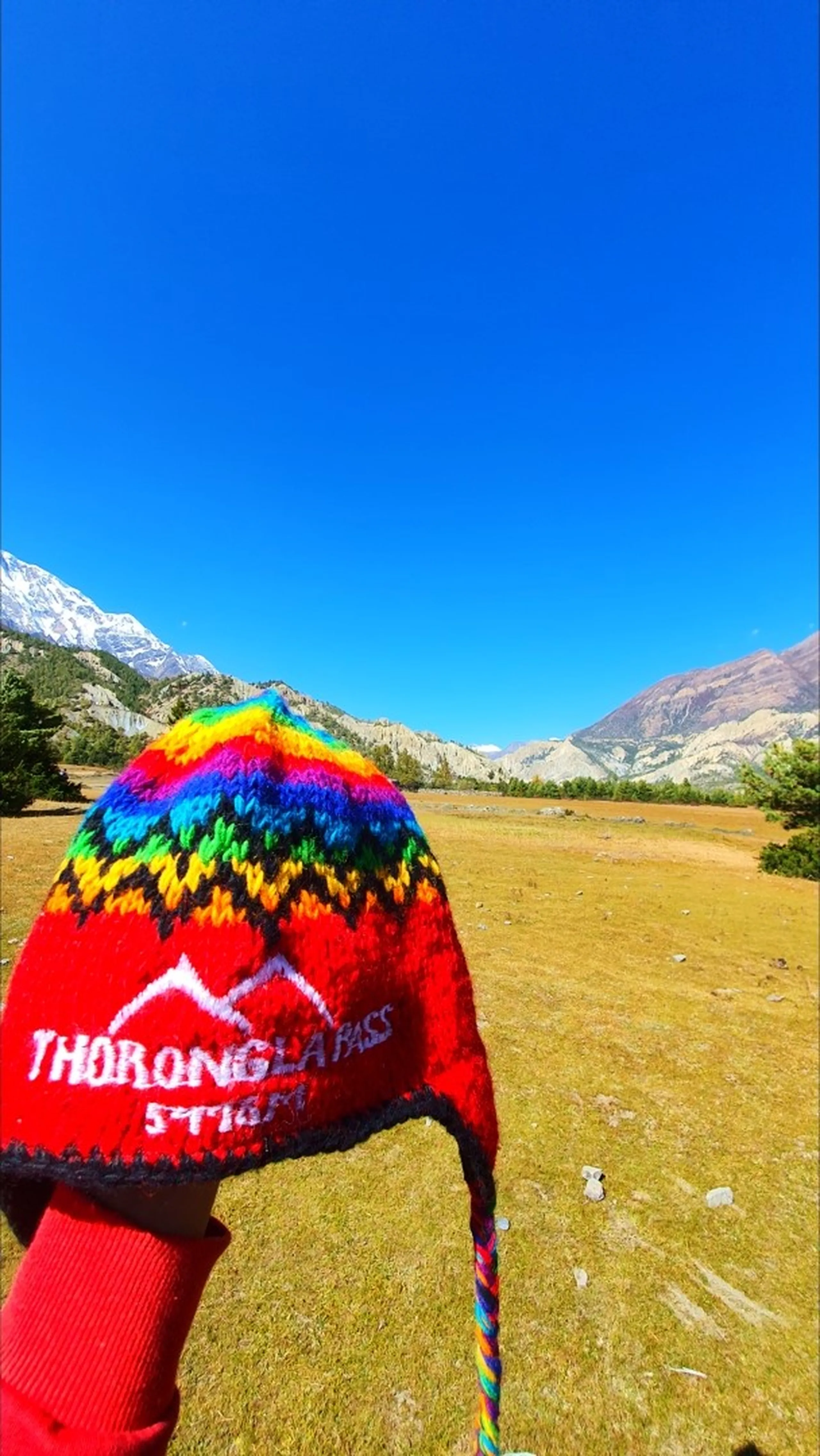 Close-up of a vibrant red hand-knitted Chullo beanie hat with rainbow-striped zigzag bands and white embroidered text reading "Thorong La Pass 5416m" held up against the wide open Manang valley, golden meadow in the middle ground, arid Himalayan cliffs and a glimpse of Annapurna snowfields on the left under vivid blue sky - Thorong La Pass Nepal souvenir hat, Annapurna Circuit trekking hat, best Nepal trek souvenir