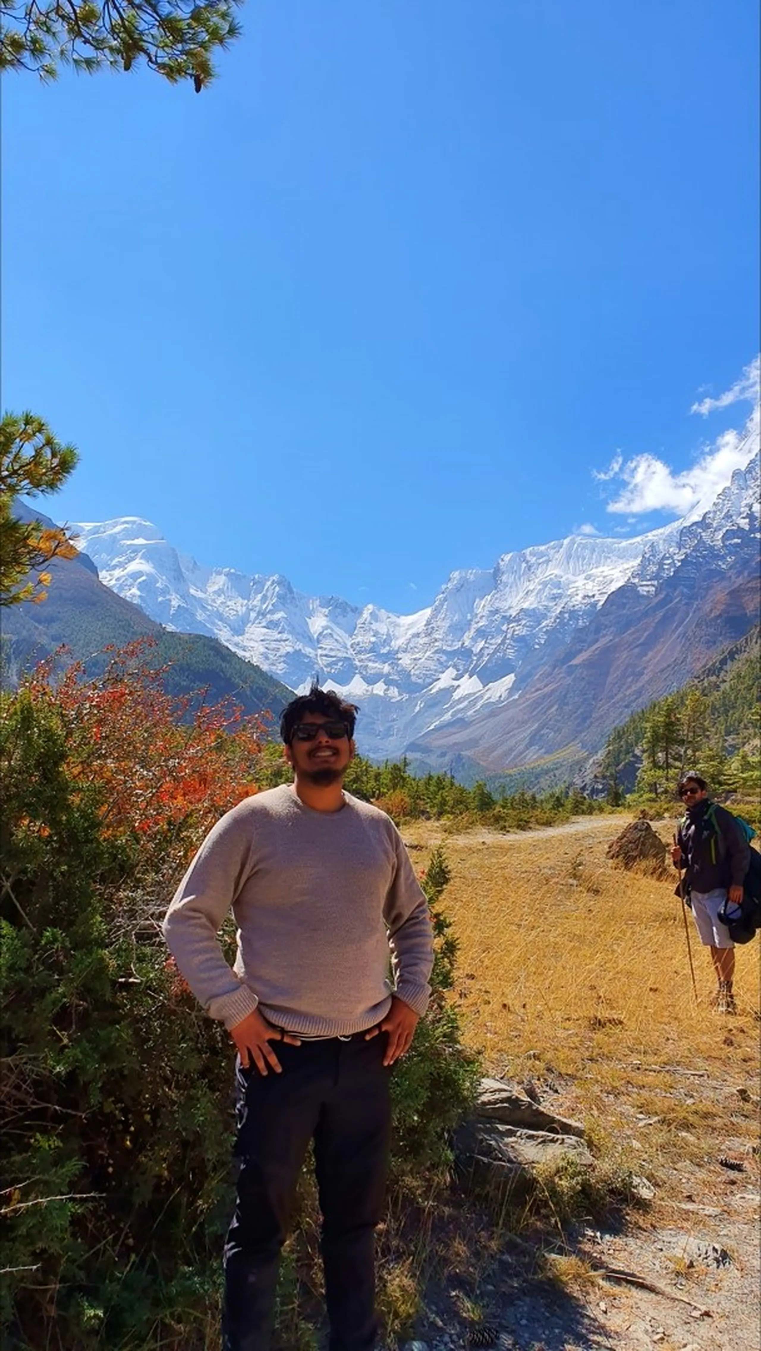 Smiling male trekker in a beige crewneck sweater and black sunglasses standing confidently with hands on hips on a rocky trail near Manang Nepal, brilliant red autumn shrubs beside him, another trekker with a large backpack visible further down the path, and the dramatic Annapurna snowfields and satellite peaks rising above the Marsyangdi valley behind him under blue sky - Annapurna Circuit Manang trail portrait, autumn trekking Nepal, Himalayan trek smile
