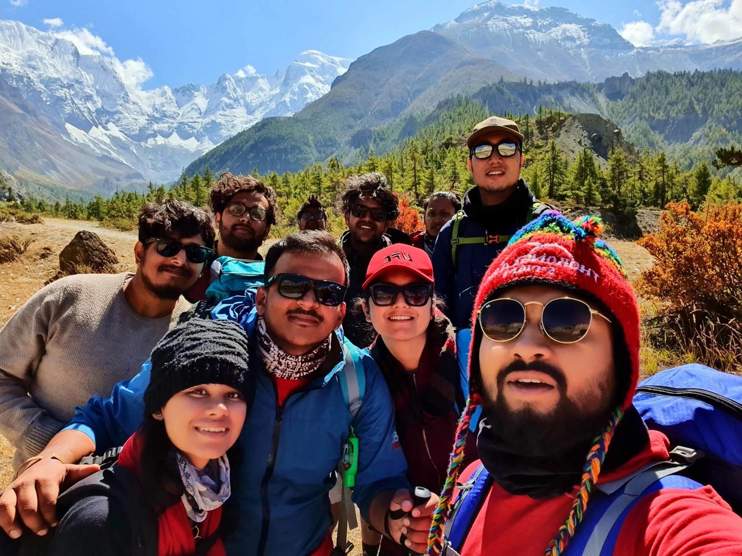 Vibrant close-up group selfie of approximately ten trekkers on the Annapurna Circuit near Manang Nepal, taken by the bearded group member in a red hoodie and colourful Thorong La Pass Chullo hat with round sunglasses in the foreground; friends crowding in behind him smiling broadly, including a woman in a red cap and dark sunglasses, another in a black beanie, and a taller member in a cap at the back - Annapurna snowfields and pine-covered valley visible behind - Annapurna Circuit group selfie Nepal, Manang trek squad, best trekking group Nepal