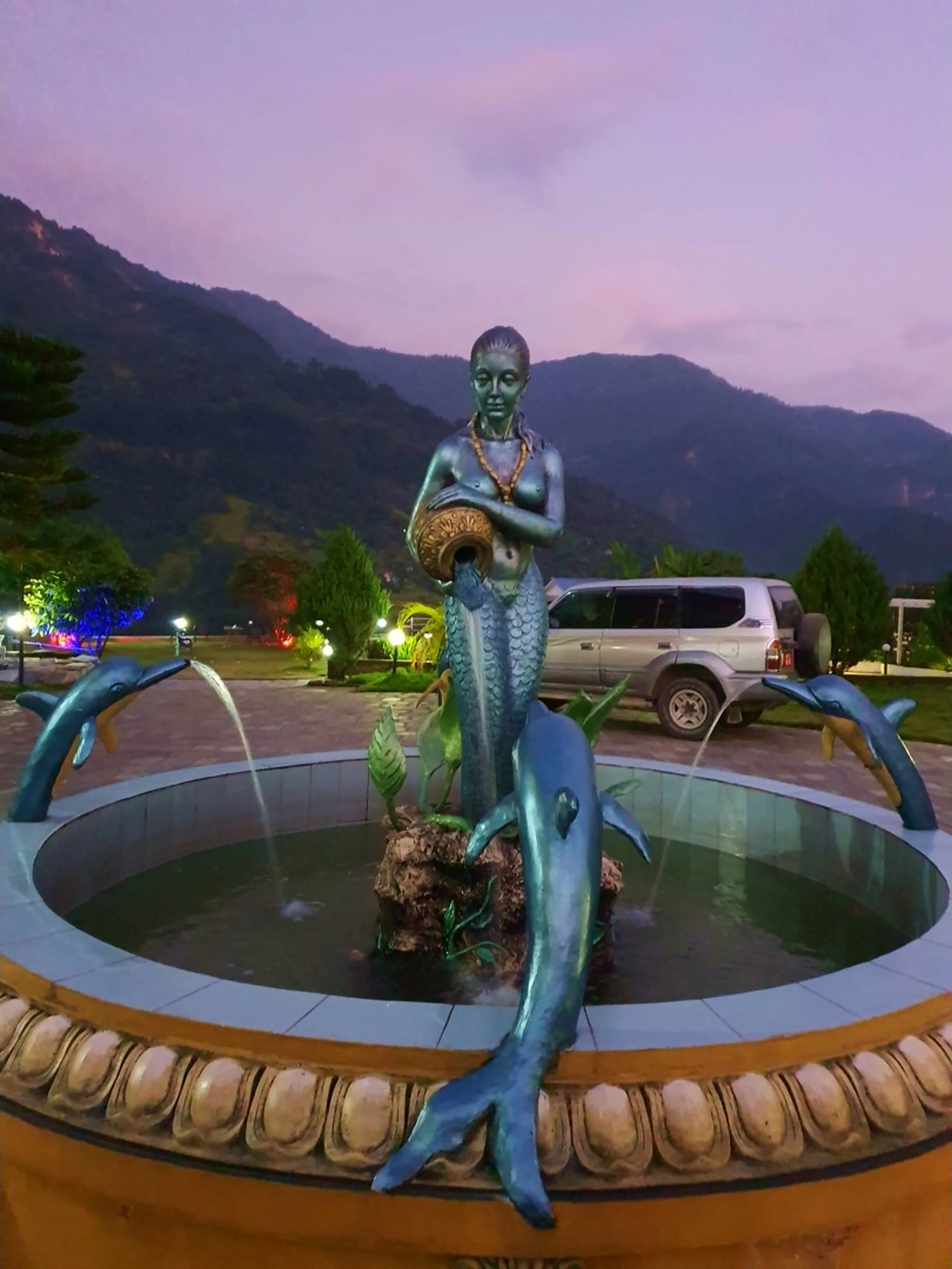 A bronze mermaid fountain sculpture pouring water from a decorative vessel, flanked by two dolphin sculptures on a circular tiled fountain basin, photographed at twilight against a dramatic purple-pink dusk sky and dark blue silhouetted Himalayan mountains - warm garden lights illuminate surrounding pine trees, a parked silver Land Cruiser visible in the background - taken at Gateway Himalaya Resort in Besisahar, Lamjung, Nepal, the ideal base camp resort for Annapurna Circuit trekkers, blending artistic sculpture with breathtaking mountain backdrop