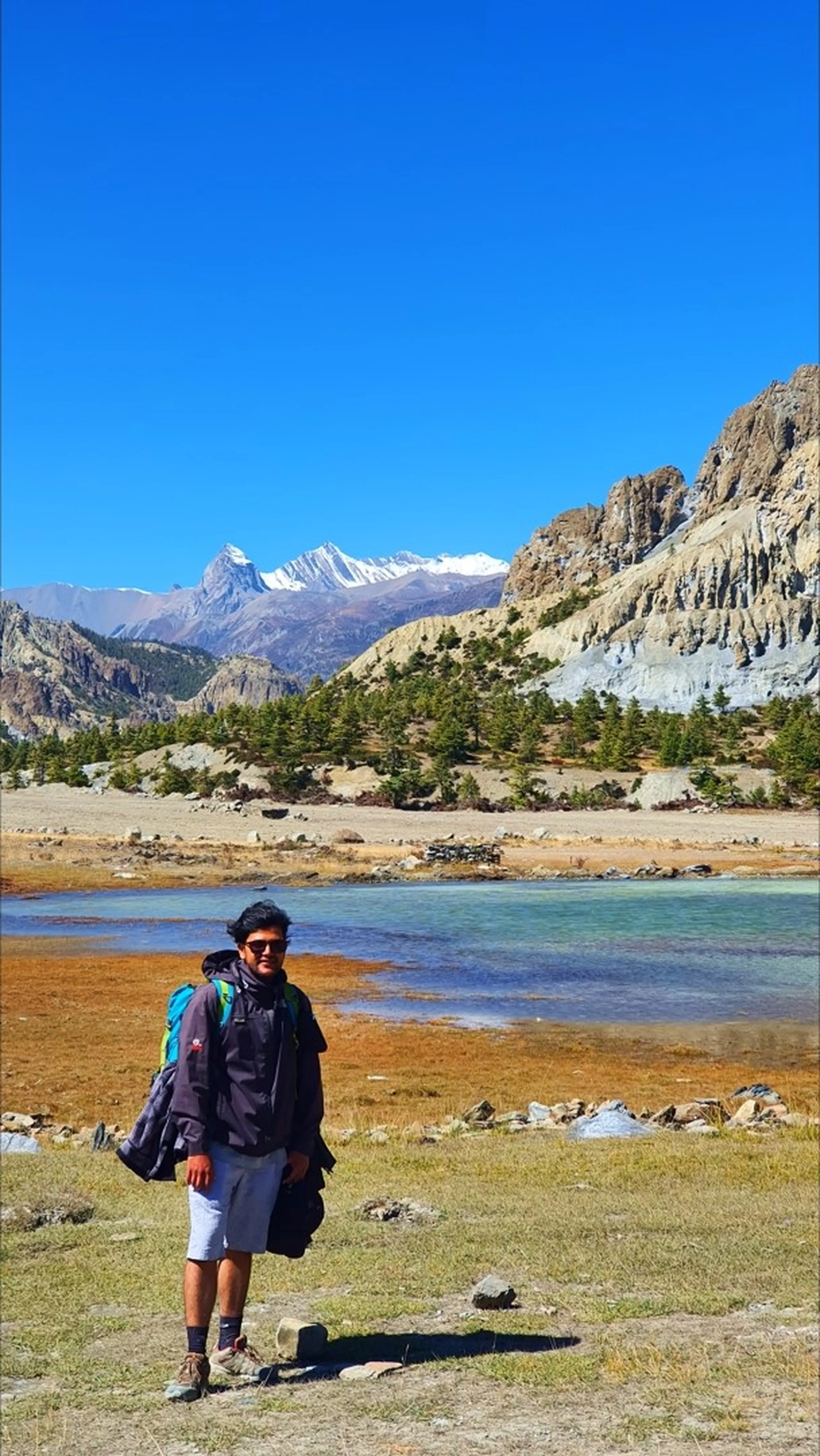 Male trekker in a dark grey jacket, grey shorts, sunglasses and trekking backpack standing beside a vivid turquoise glacial lake near Manang Nepal, surrounded by eroded sandy ochre rock pinnacles and spires on the right, sparse pine trees across the lake, and snow-capped Himalayan peaks visible in the distance under a clear blue sky - glacial lake Manang Nepal, Annapurna Circuit scenic stop, high altitude lake Nepal trekking